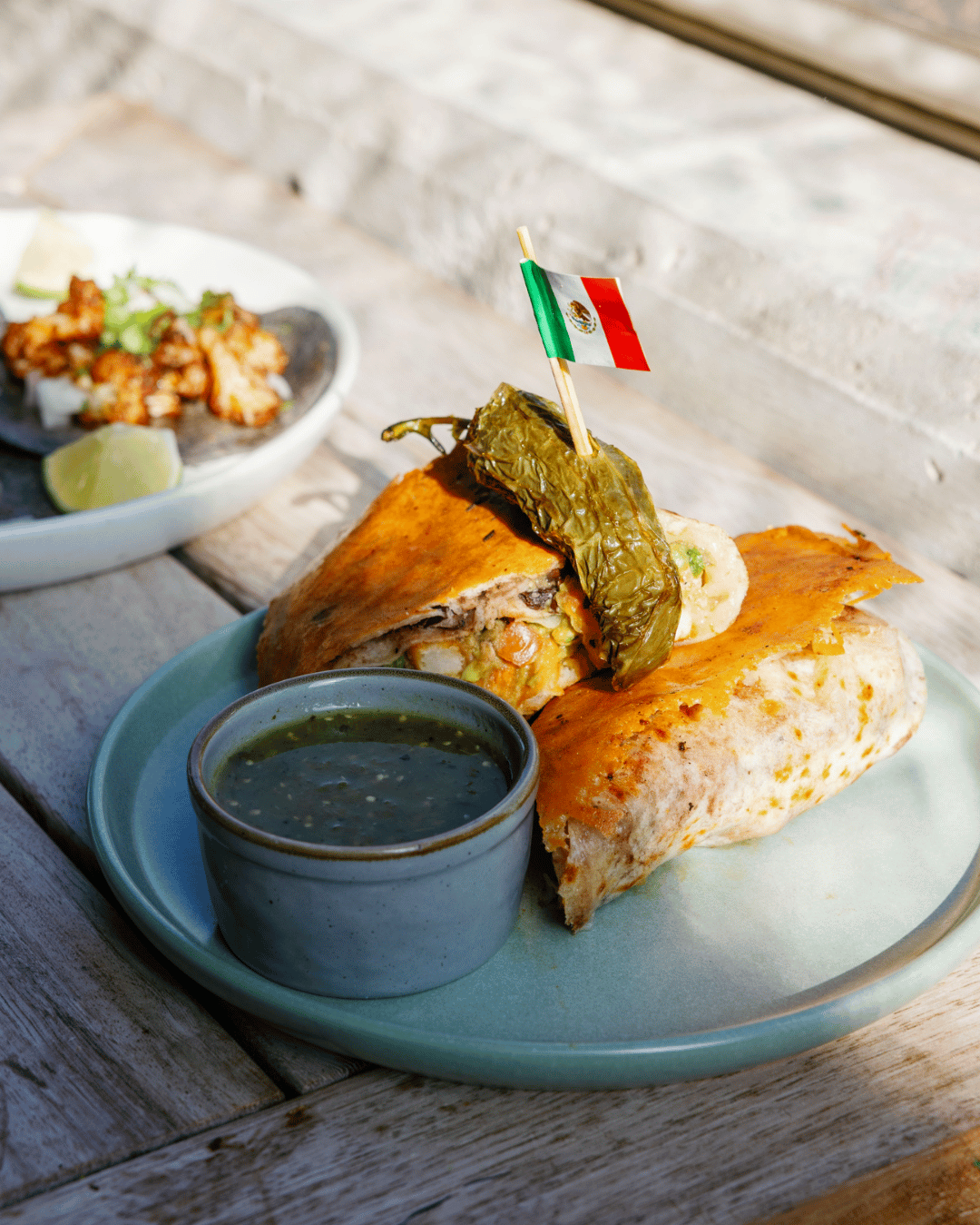 A plate with a burrito topped with a pickled pepper and a small Mexican flag, served with green salsa and lime wedges on a wooden table, with a side dish of fried seafood in the background.
