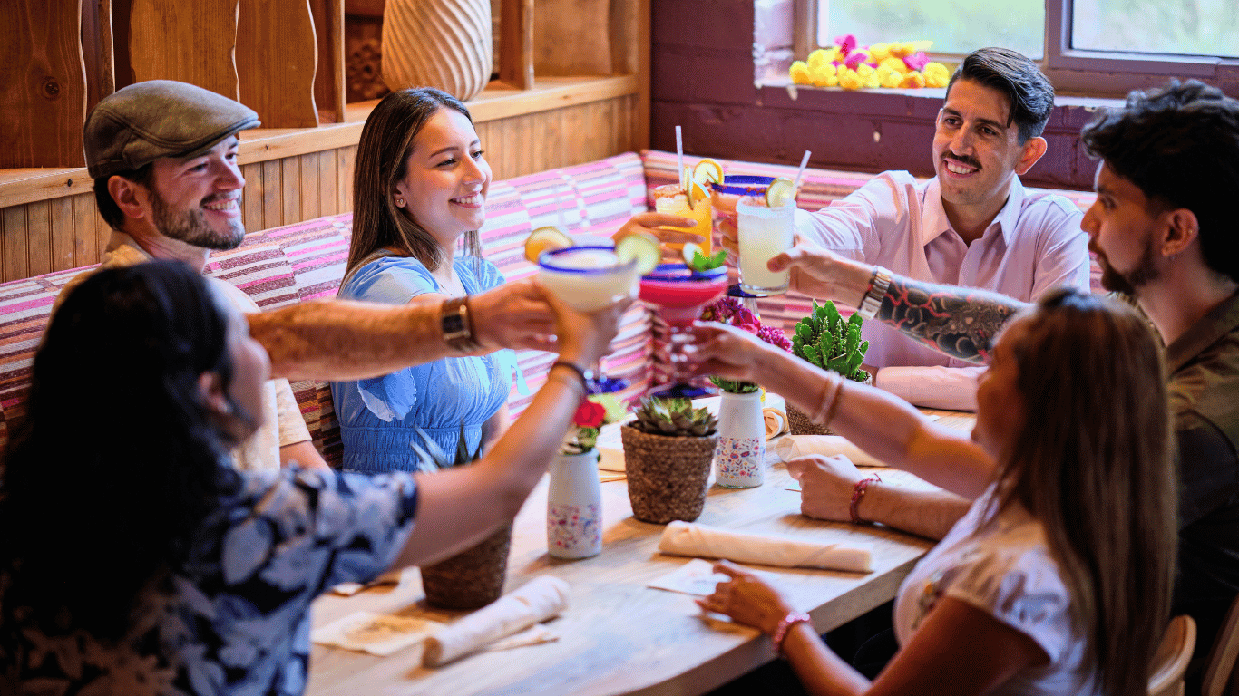 Group of six friends celebrating with drinks at a restaurant table, clinking glasses, smiling, and enjoying each other's company in a cozy, rustic setting with wooden walls and colorful decorations.