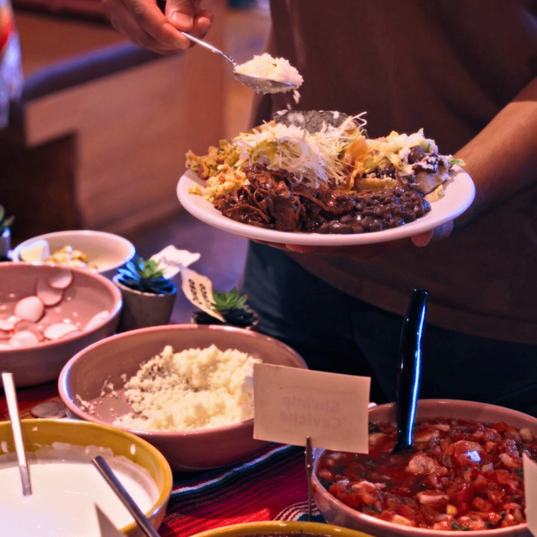 Person serving food onto a plate containing shredded lettuce, pulled meat, and other traditional Mexican dishes. Several bowls of Mexican food, including rice, beans, and salsa, are on the table.