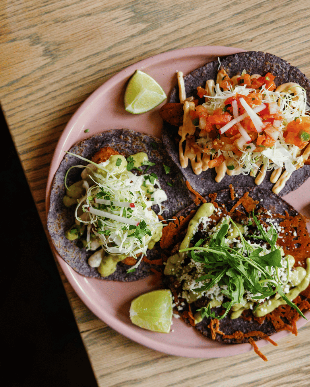 Three vibrant street tacos with various toppings on a pink plate, garnished with lime wedges, on a wooden table.