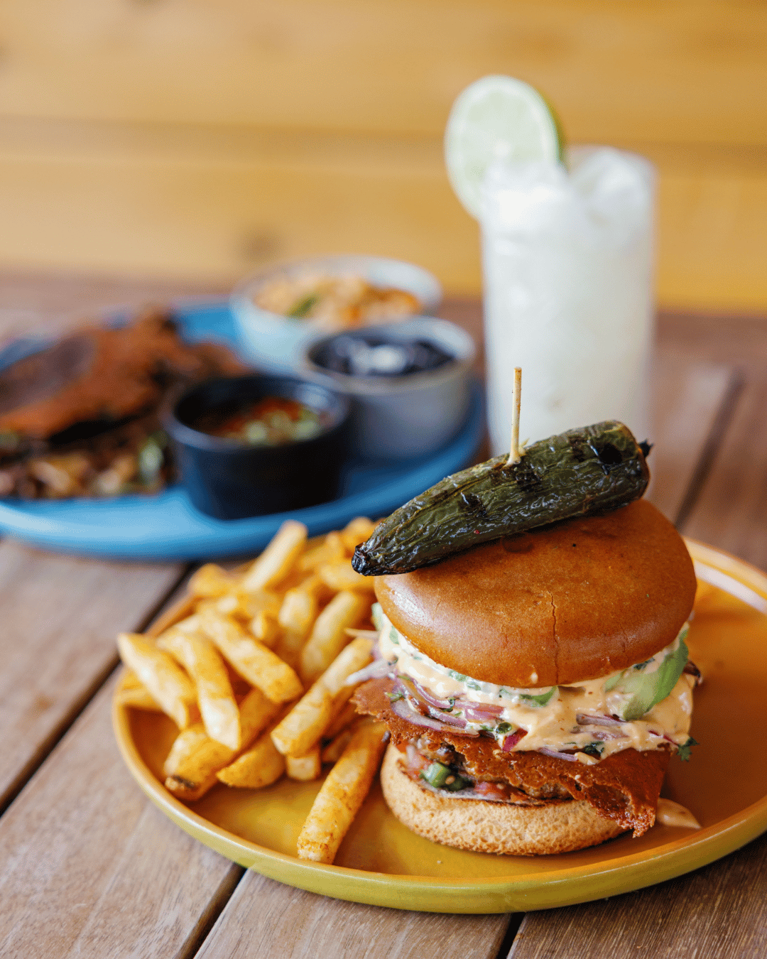 Fried chicken sandwich with lettuce and sauce, topped with a pickled jalapeño, served with French fries on a yellow plate. In the background, there is a drink with a lime wedge and a side of condiments and other food items on a blue plate.