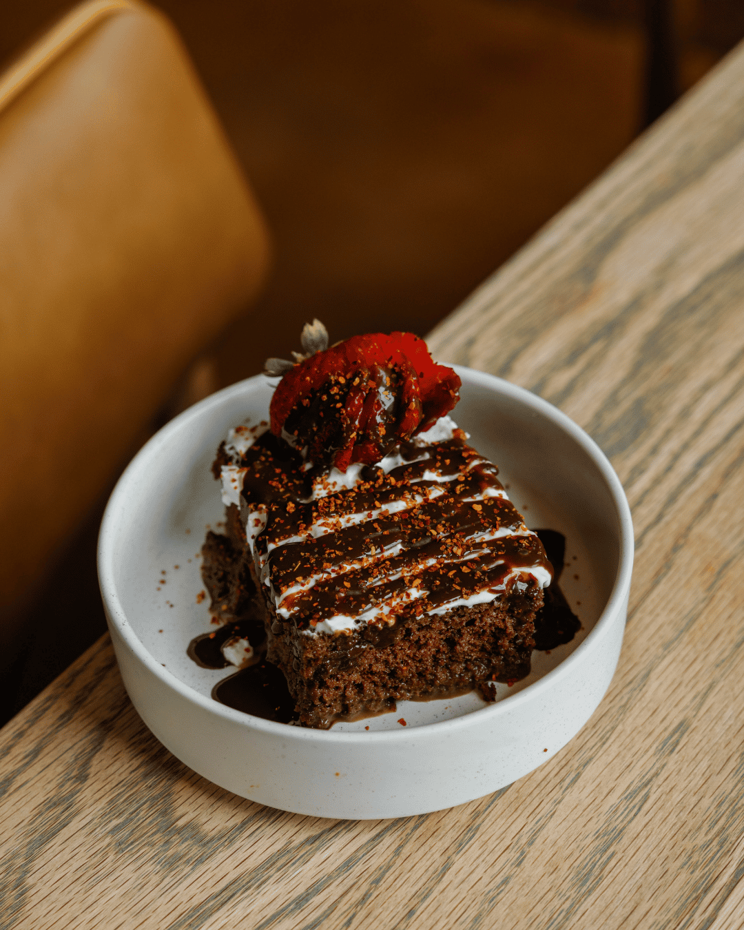 Chocolate cake topped with whipped cream, drizzled with chocolate sauce, sprinkled with crushed nuts, and garnished with a strawberry on a white plate.