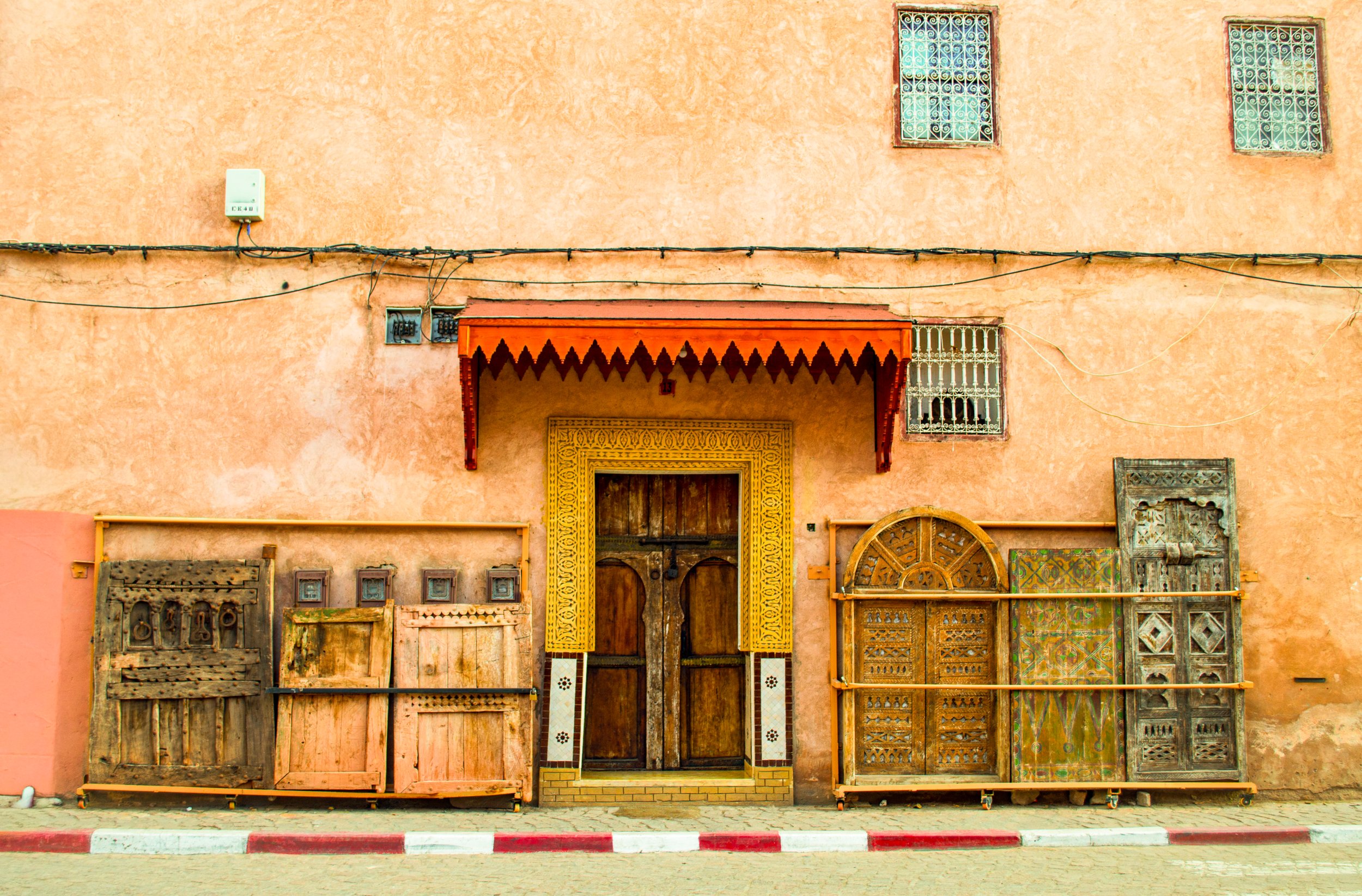 A peach-colored building with wooden doors, decorative metal window guards, and a red awning. The facade has various old wooden and metal items displayed, creating a rustic appearance.