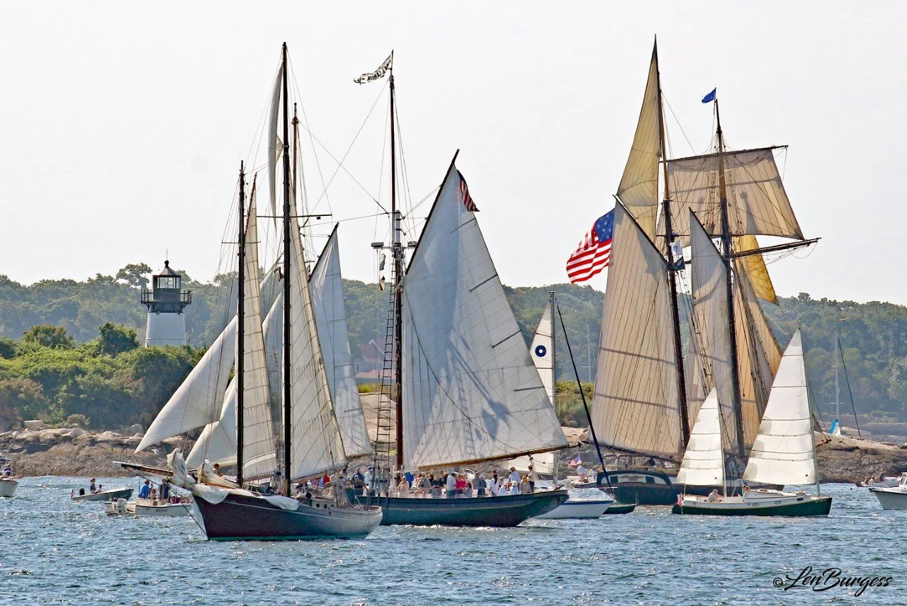 Several sailboats with white and beige sails are sailing on the water near a rocky shoreline in Gloucester, MA with a lighthouse in the background and a wooded hillside with a lighthouse on top.