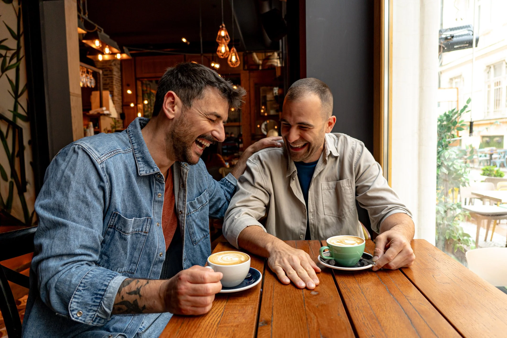 Two men are sitting at a wooden table in a cozy café, laughing and enjoying each other's company with cups of coffee in front of them. The café has warm lighting and a large window letting in natural light.