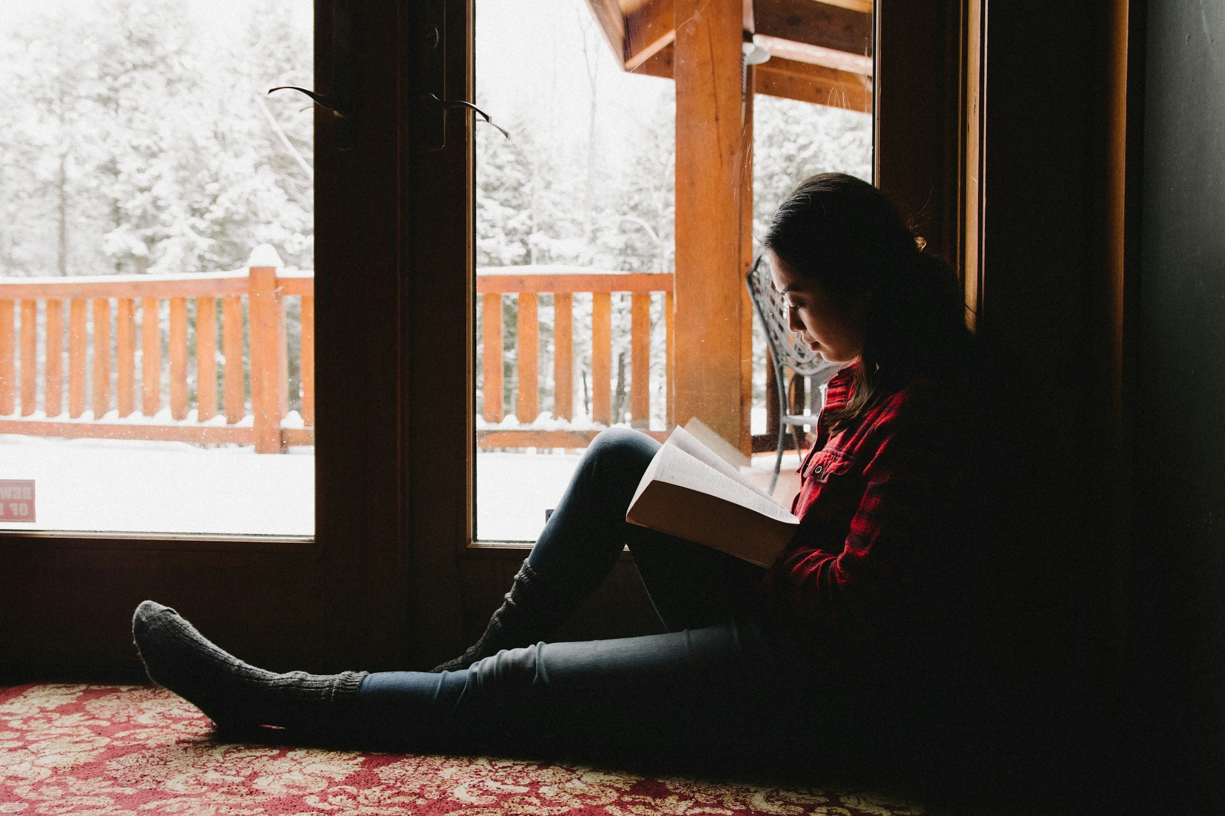 A young woman with long dark hair, glasses, and a red plaid shirt sitting by a window, reading a book. Outside the window, snow covers the ground and trees, with a wooden deck railing visible.