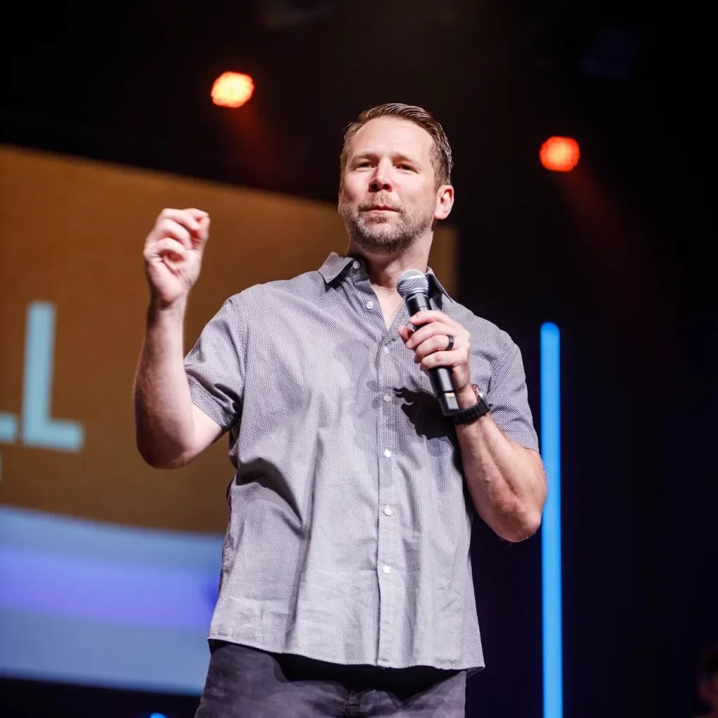 A man with a beard holding a microphone on stage, looking confident, wearing a short-sleeved, button-up shirt, with a blurred background and stage lights.