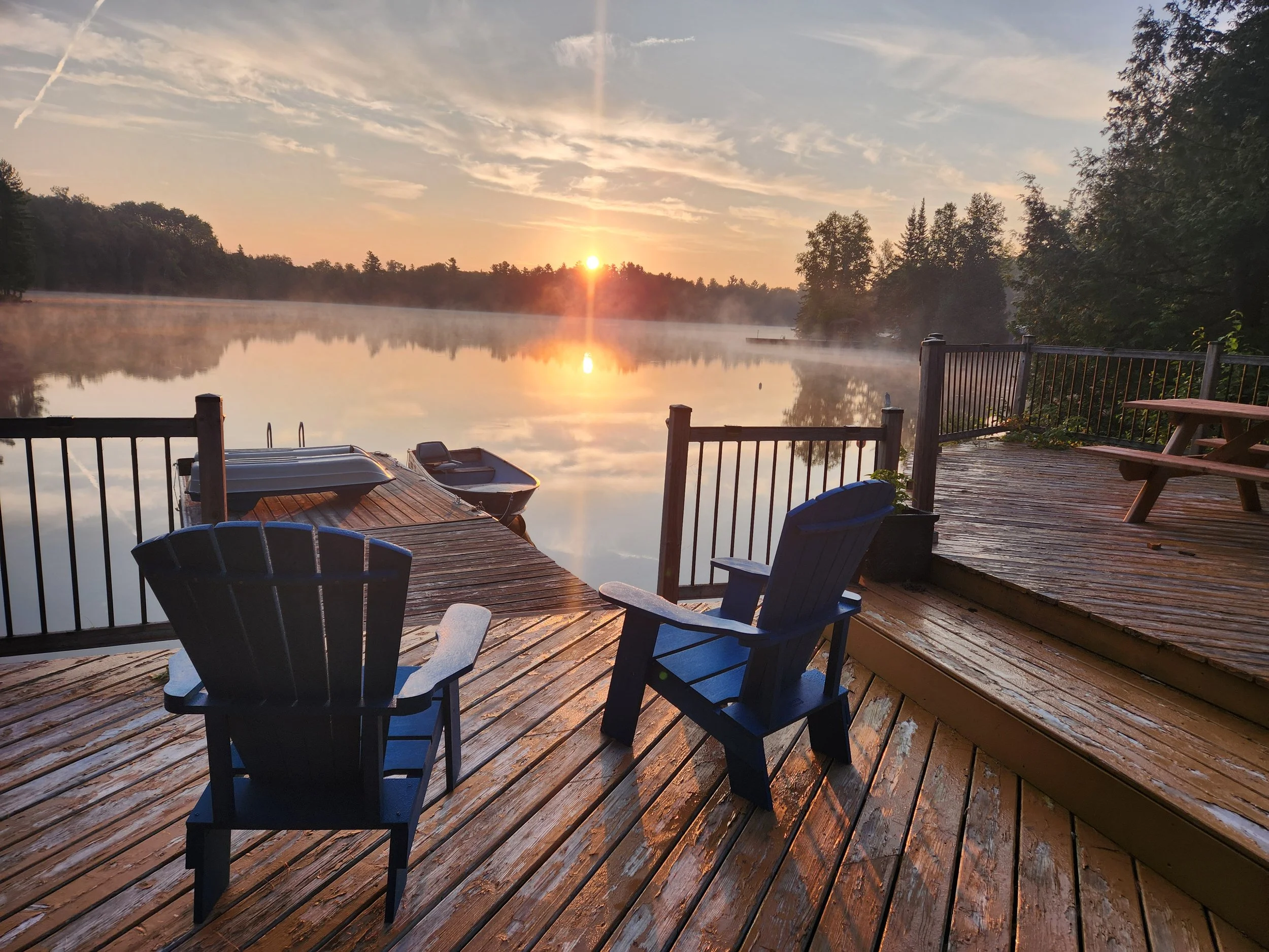 A lakeside wooden deck at sunrise with two blue Adirondack chairs, small boats tied to the dock, and mist rising on the water. Trees border the lake, and the sky has light clouds with the sun on the horizon.