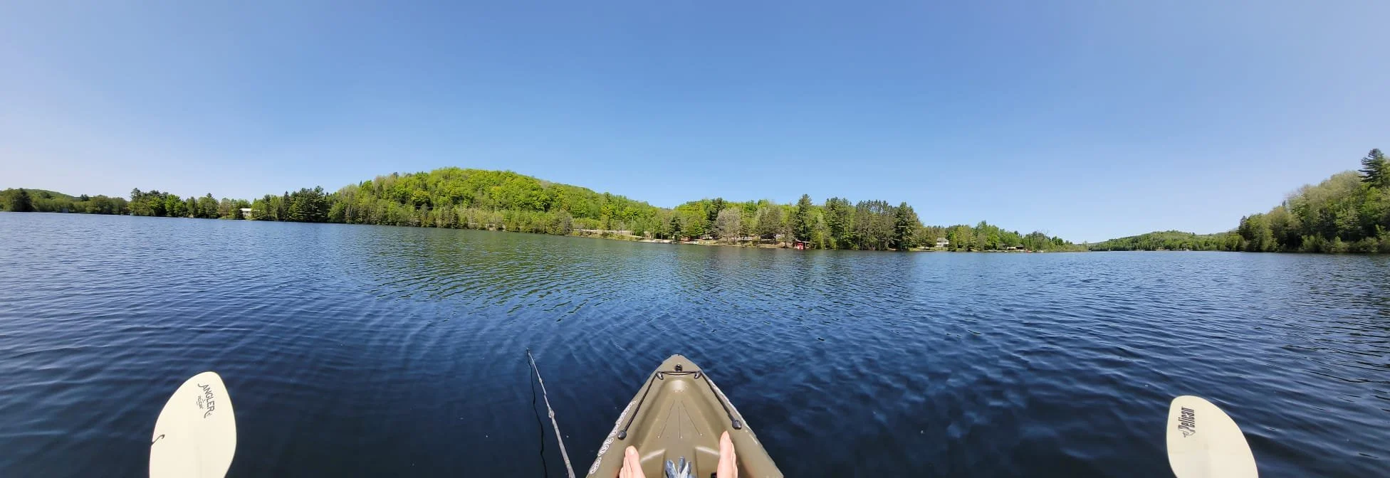 View from a kayak on a peaceful lake with green trees and hills in the background under a clear blue sky.