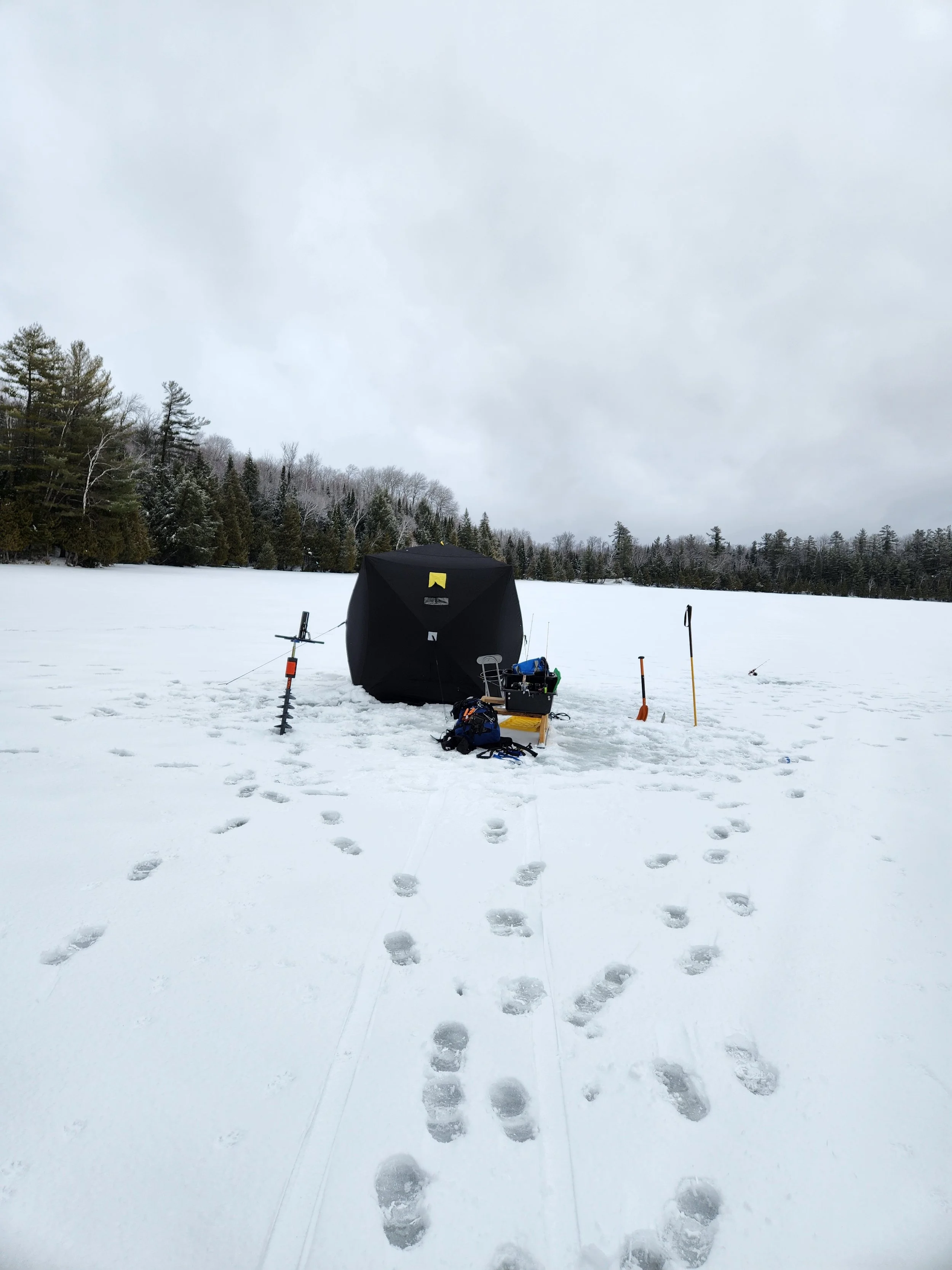 Ice fishing setup on a snow-covered lake with a shelter, tools, and equipment, surrounded by trees under a cloudy sky.