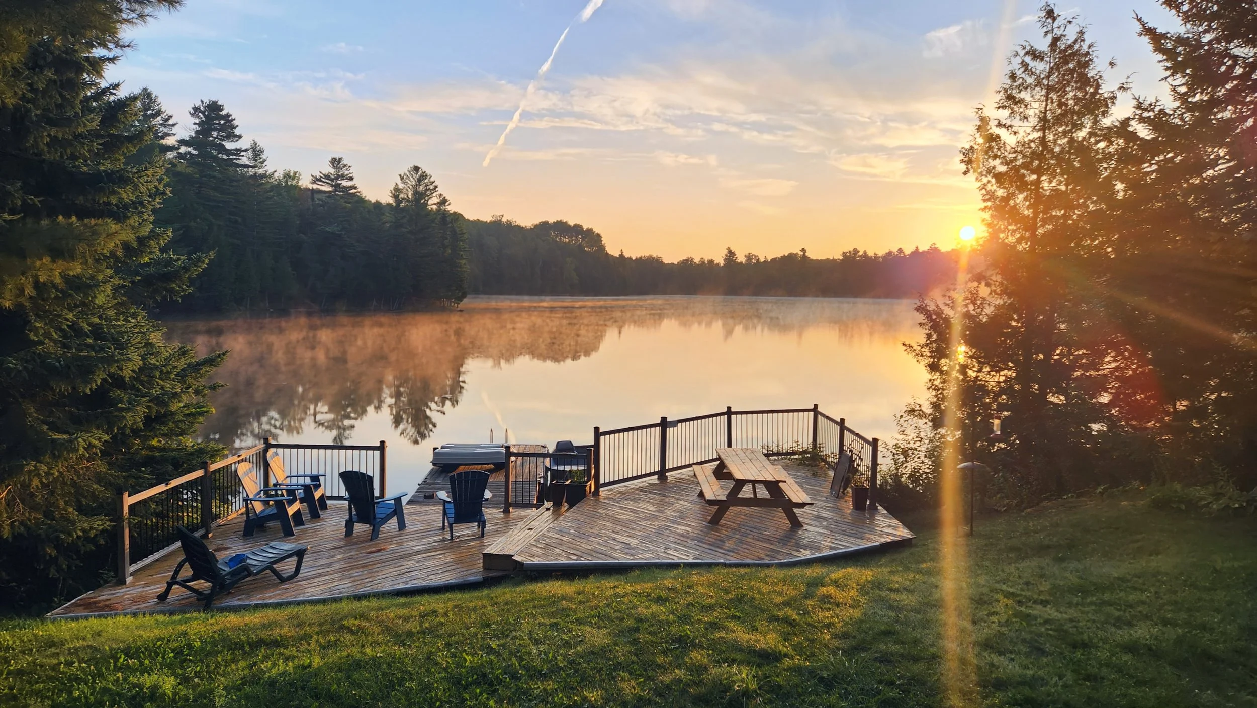 A wooden deck with chairs, a picnic table, and a small hot tub, overlooking a calm lake at sunrise with mist rising from the water and trees on the far shore.