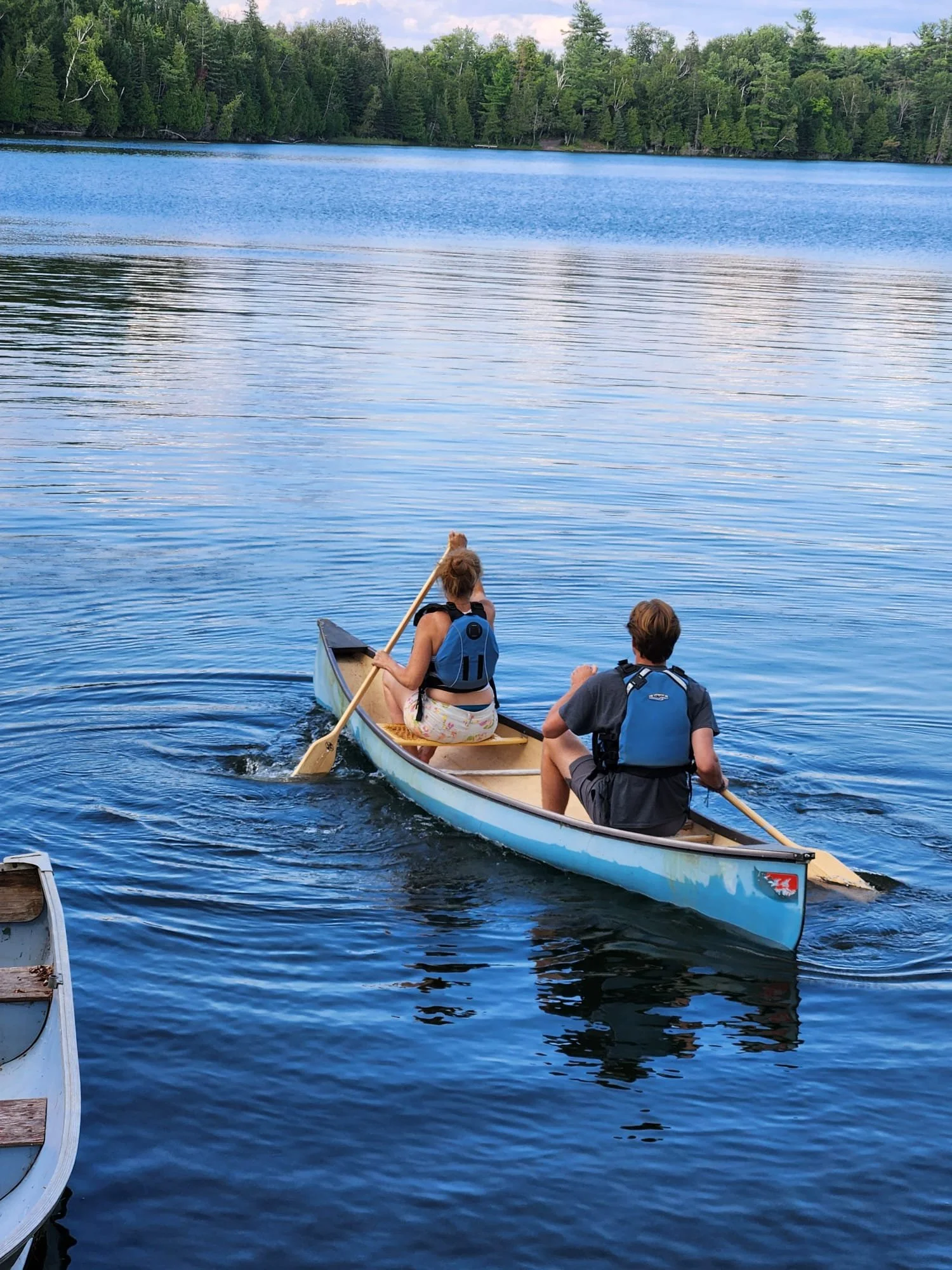 Two people paddling a canoe on a calm lake with a forested shoreline in the background.