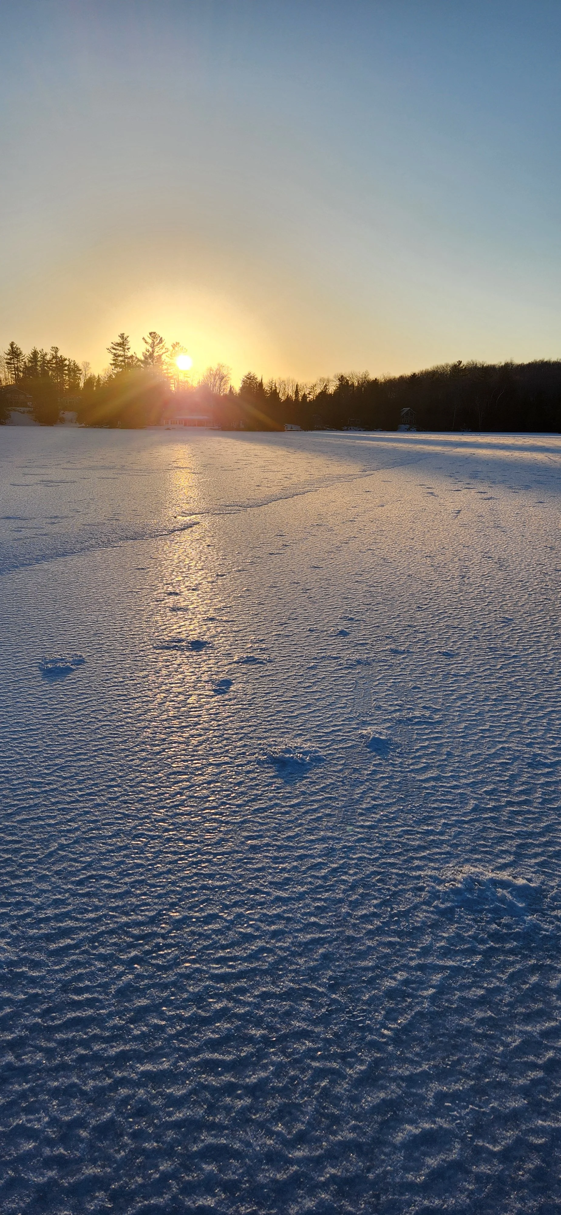 Snow-covered frozen lake at sunset with footprints in the snow and trees in the background.