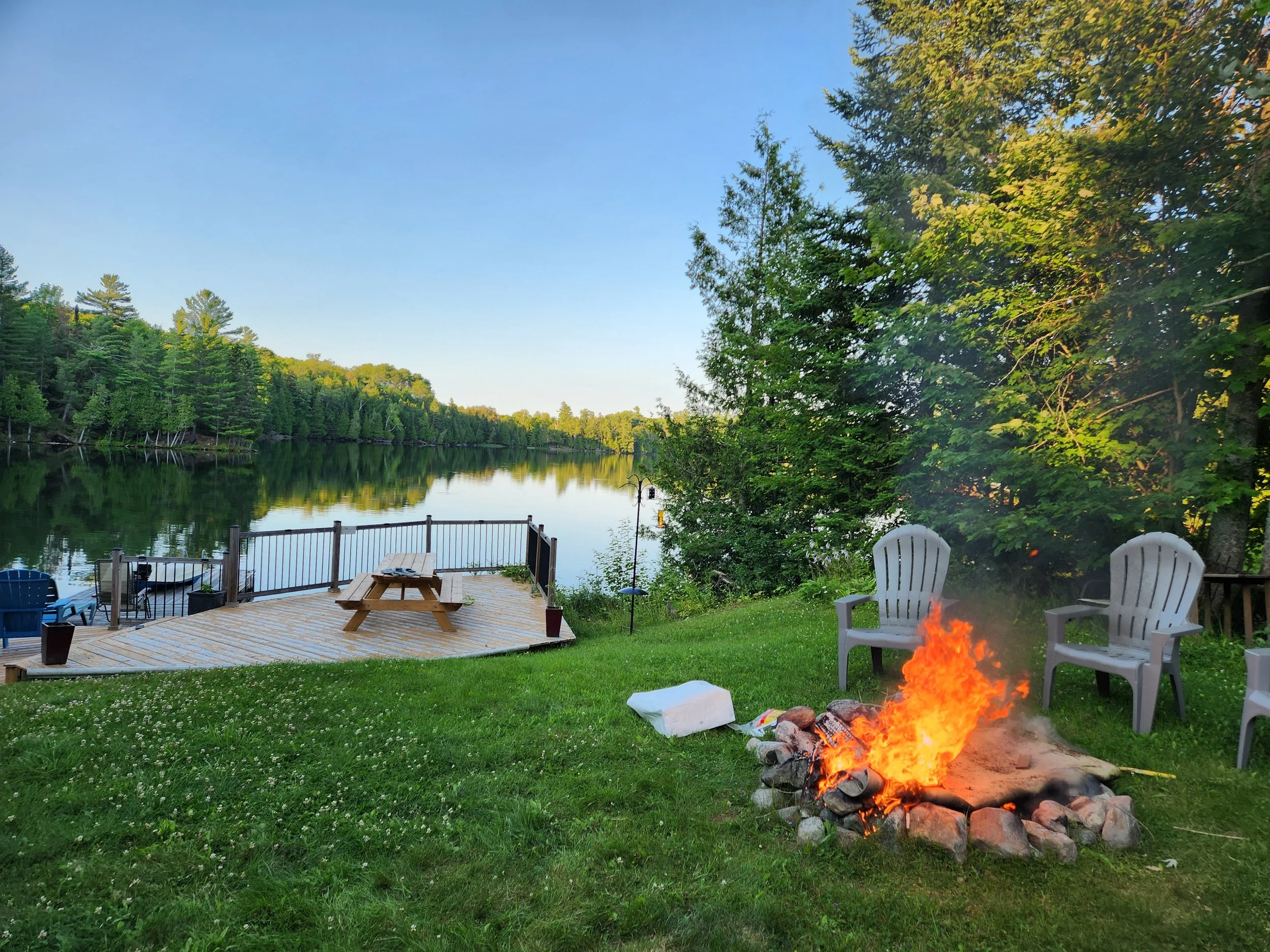 A lakeside backyard with a fire pit, white Adirondack chairs, a wooden deck with a picnic table, and a view of a calm lake surrounded by trees.