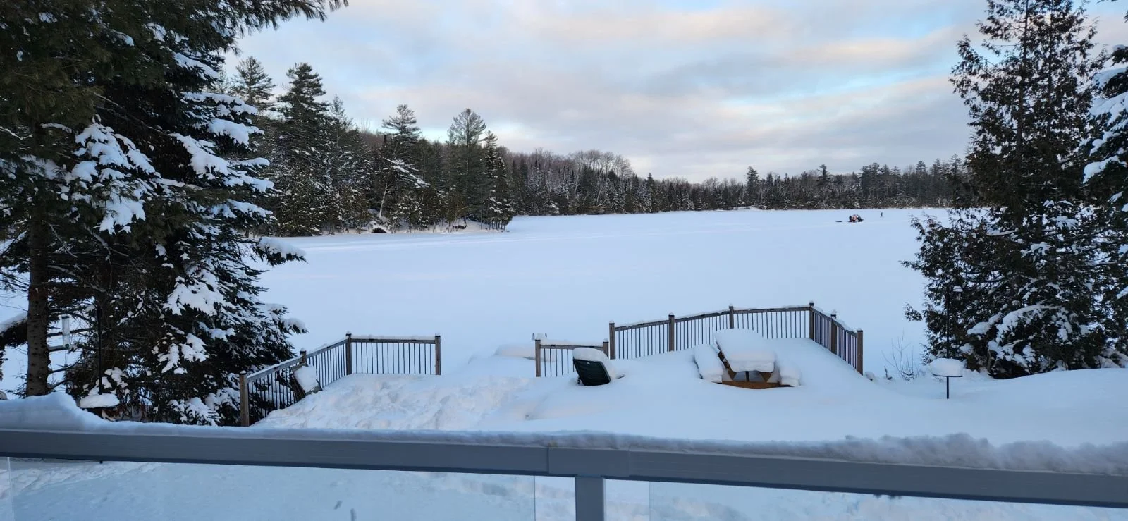 Snow-covered backyard with a wooden deck and trees, looking out over a frozen lake with a snow-covered shoreline, in a winter landscape.