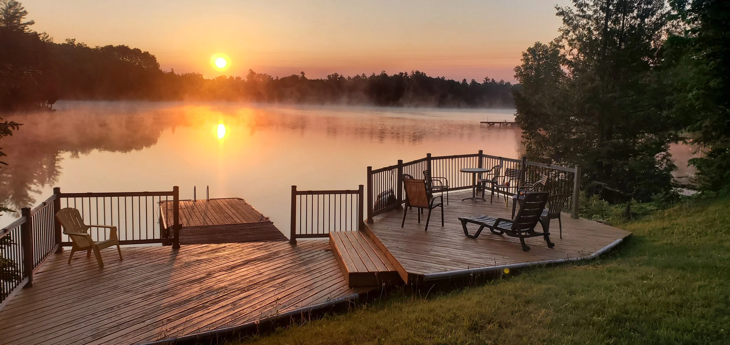 Sunrise over a calm lake with a wooden deck and outdoor seating, surrounded by trees and mist rising off the water.