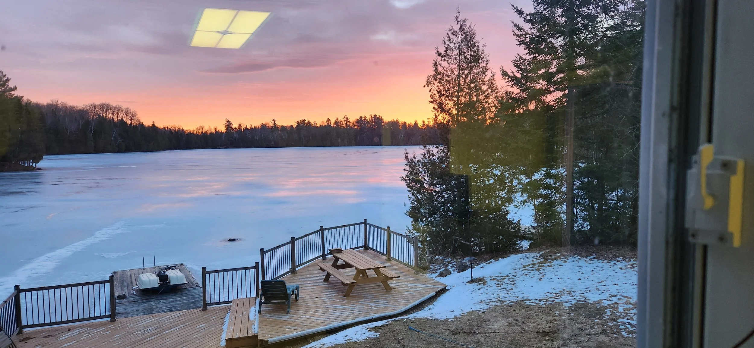 A view of a frozen lake with a snow-covered shoreline, a wooden deck with a picnic table and a chair, surrounded by trees, during sunset.