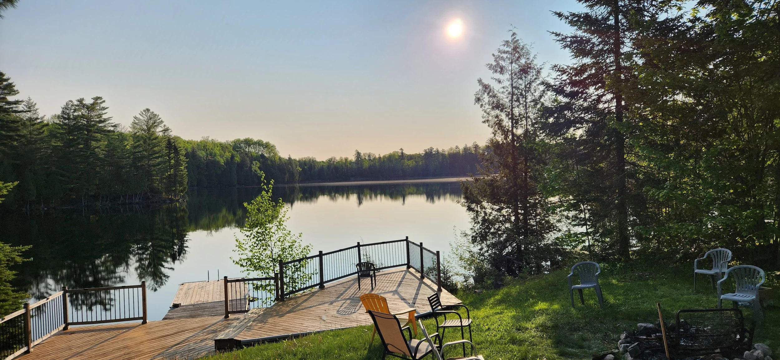 A lakeside scene with a wooden dock and a grassy area with chairs and trees, reflected in the calm water under a bright sun.