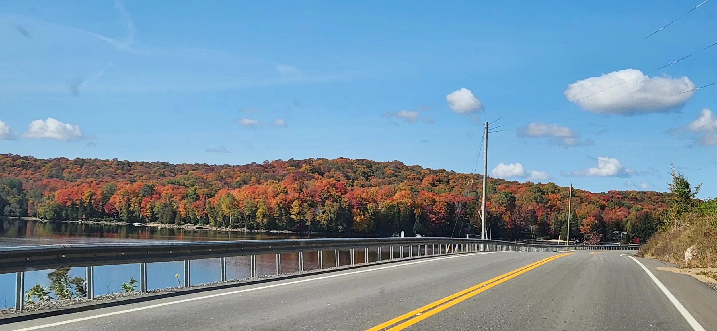 A scenic road during fall with colorful trees on a hillside and a body of water beside the road, under a bright blue sky with scattered clouds.