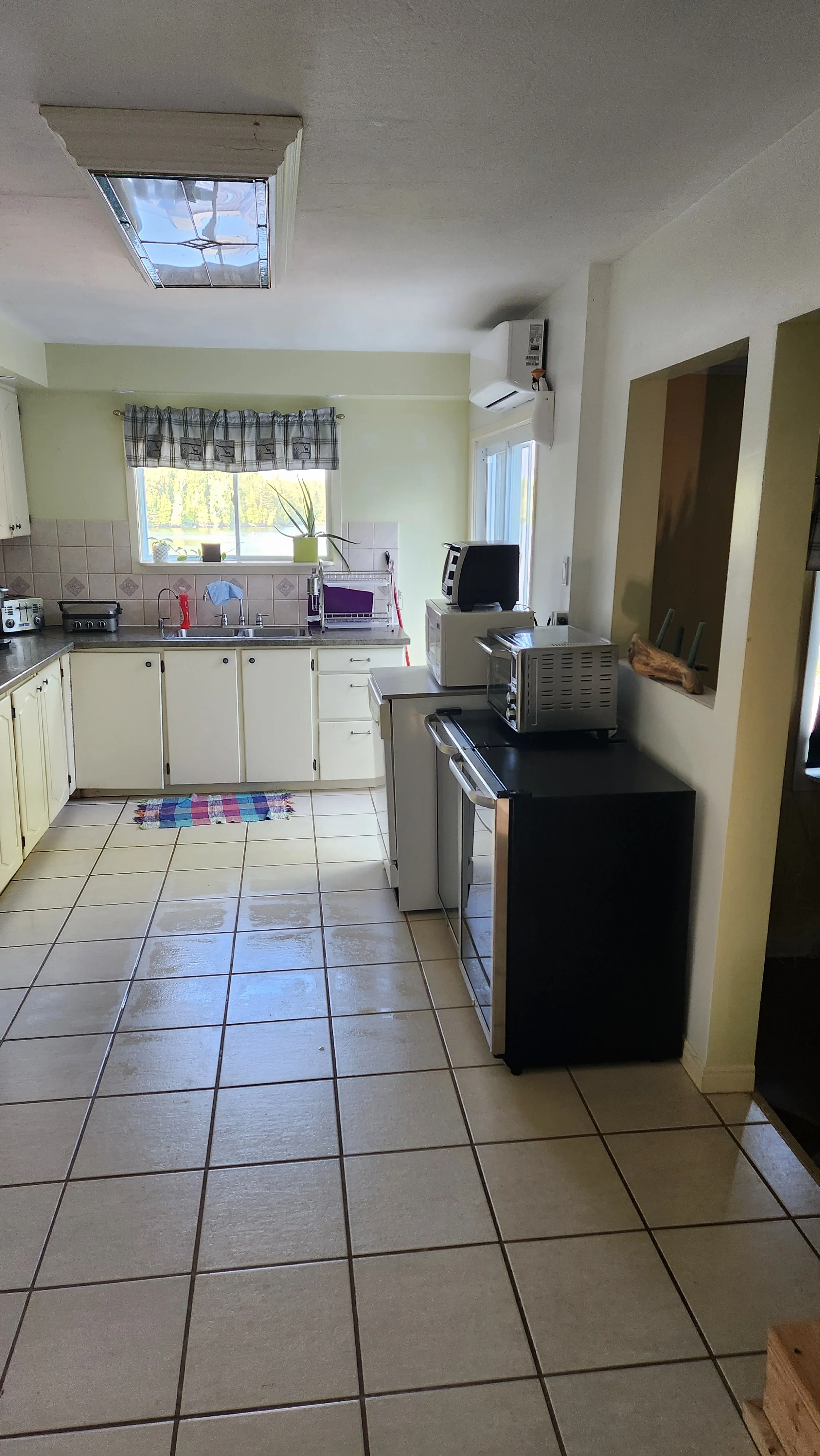 A kitchen with cream-colored cabinets, beige tiled floor, a window with a plaid valance, and various appliances on the counters including a toaster, microwave, and small oven. There is a colorful rug in front of the sink and a potted plant on the windowsill.