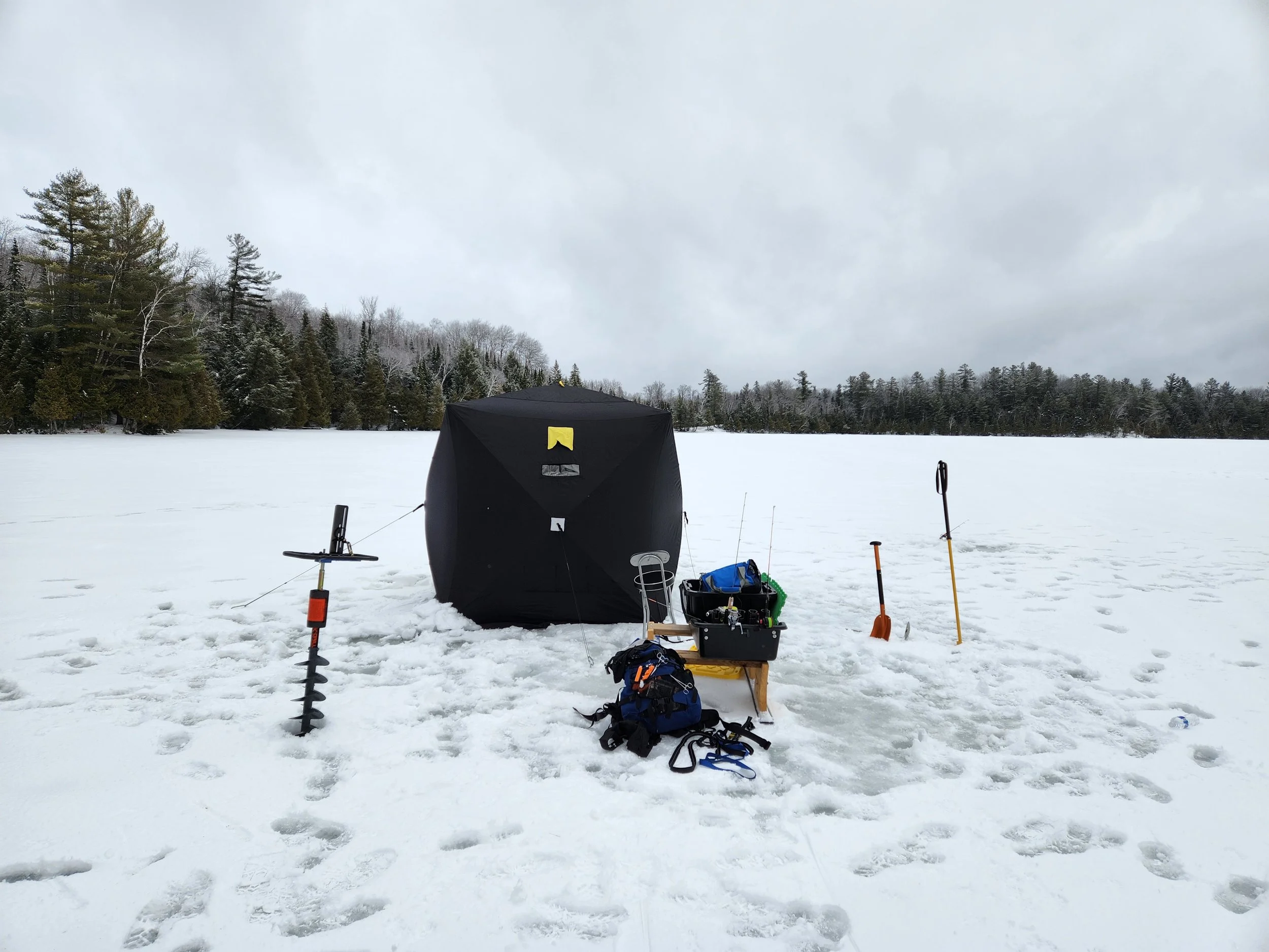 Ice fishing setup on a frozen lake with a small black tent, shovels, equipment box, and fishing gear.