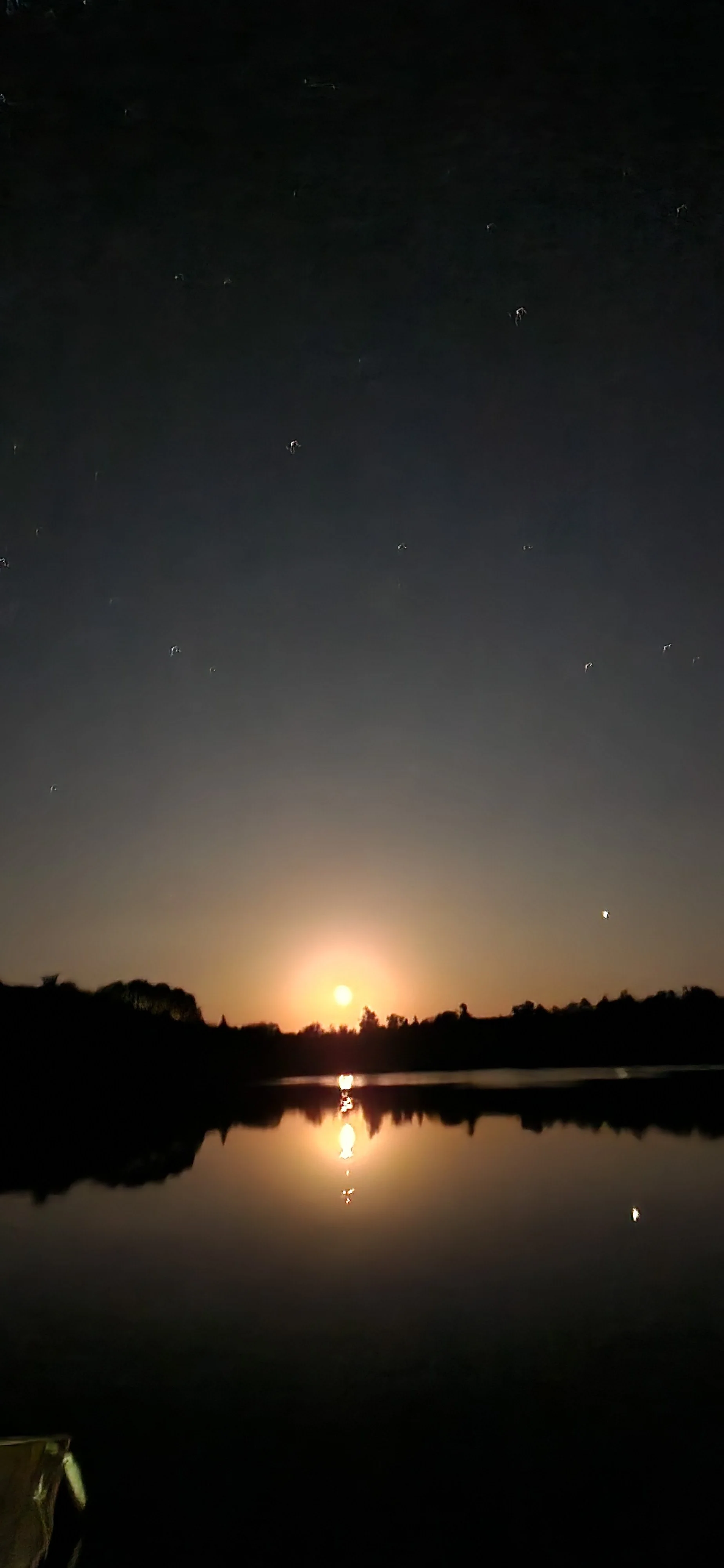 Nighttime scene of a river with a bright full moon reflecting on the water, silhouette of trees along the shoreline, and stars scattered across the sky.