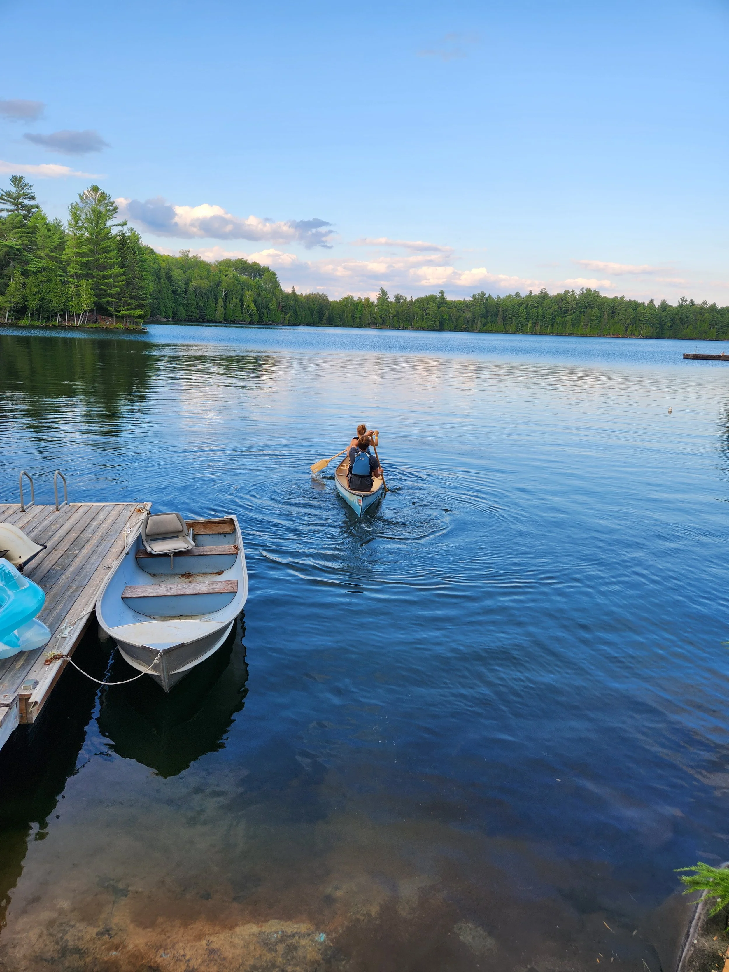 A person paddling a canoe on a calm lake surrounded by green trees, with a wooden dock and small boat in the foreground and a blue sky with clouds overhead.
