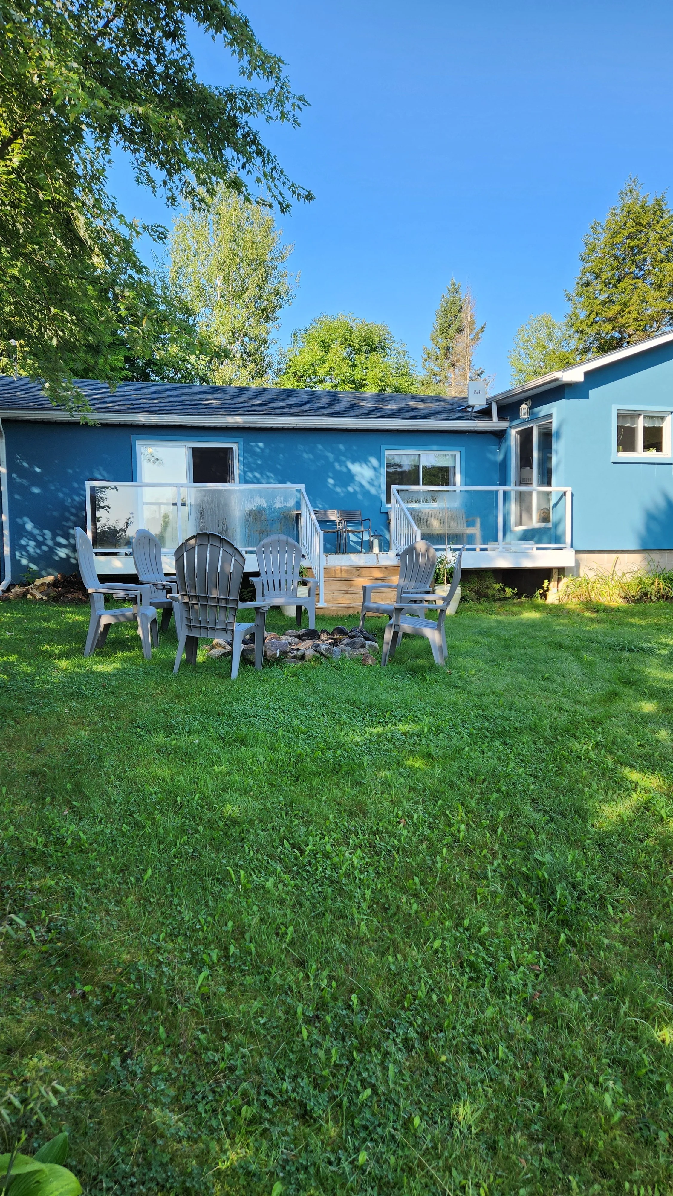 A backyard with a green lawn, a blue house with a deck, white railing, and outdoor seating, including chairs around a fire pit, and trees under a clear blue sky.