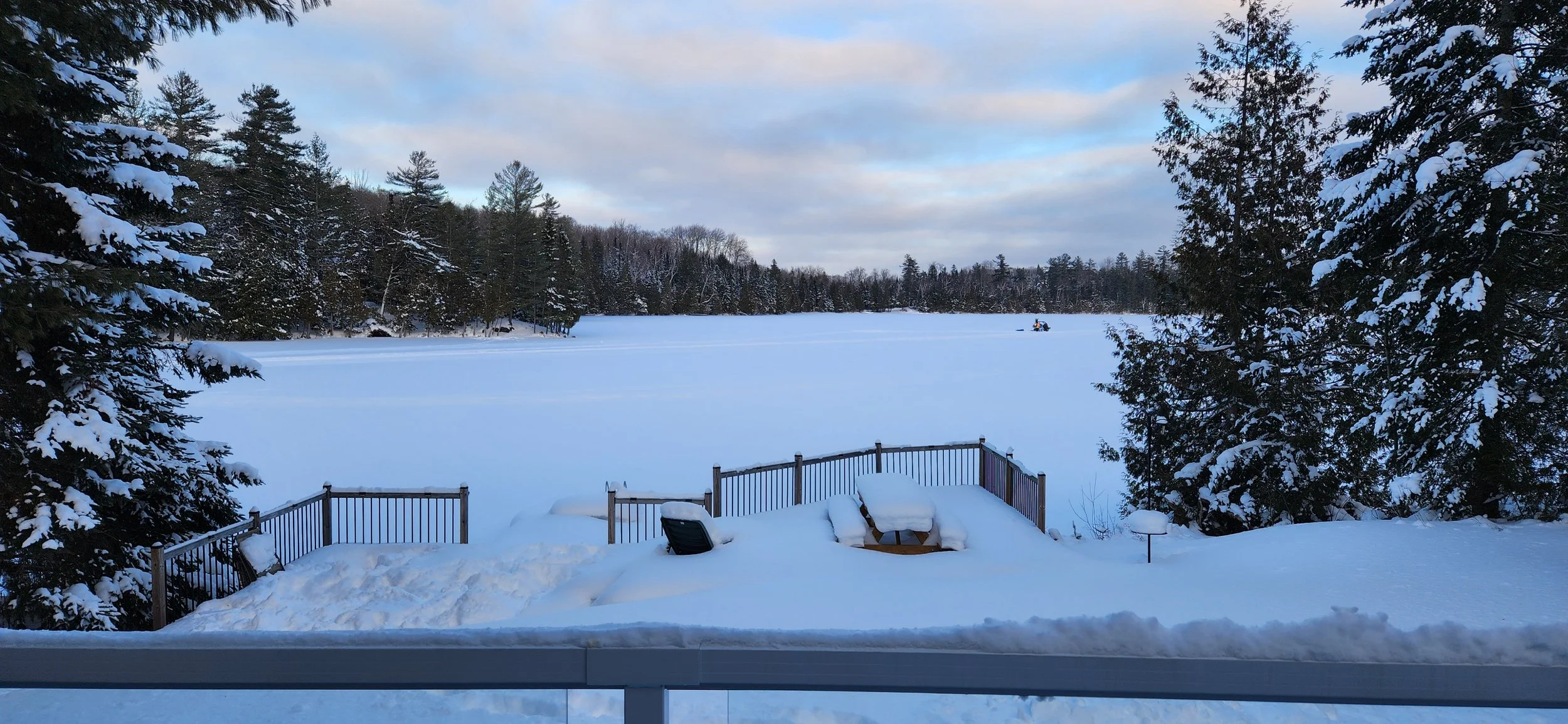 Snow-covered backyard with a deck, snow-covered furniture, and trees surrounding a frozen lake or pond in the background, under a partly cloudy sky.