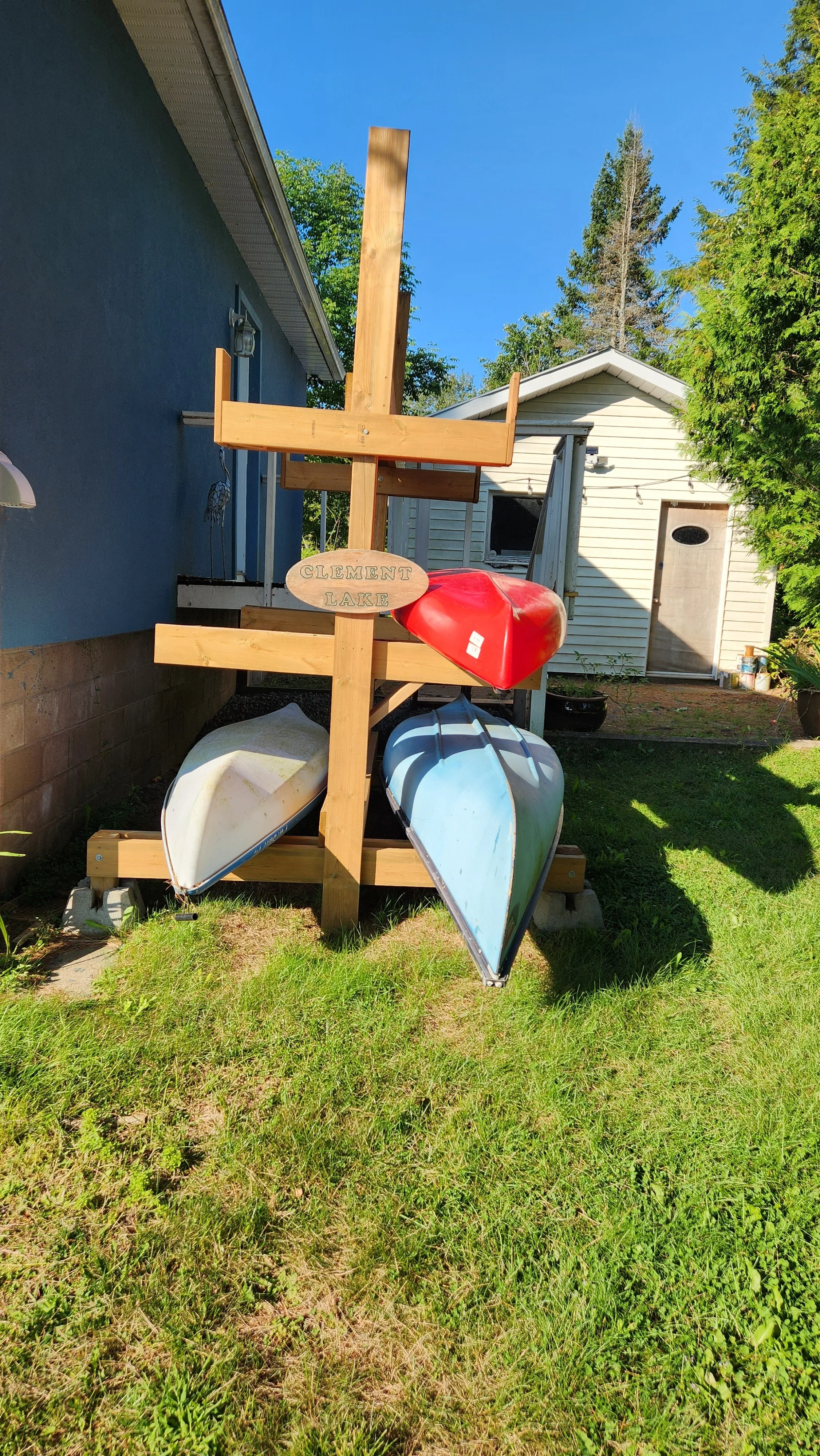 A wooden kayak rack holding three kayaks, with a sign that says "Clement Lake." Two of the kayaks are blue and white, and one is red. The rack is outside next to a house with a blue exterior and a white shed in the background, on a grassy area.