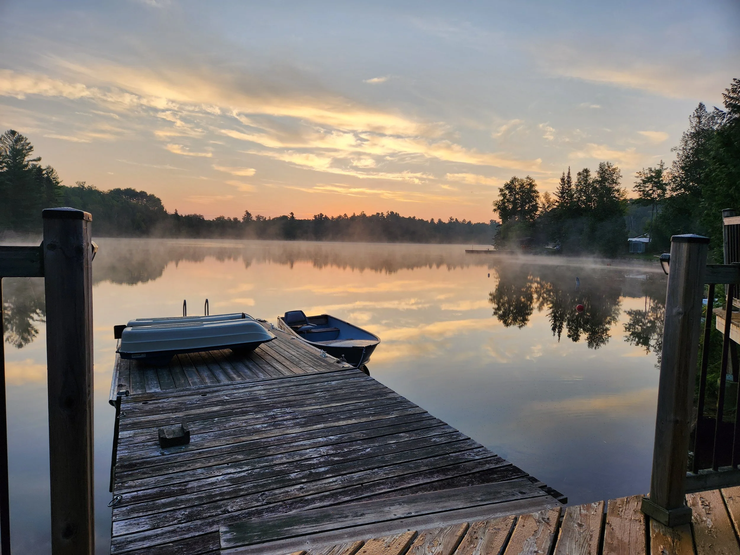 A wooden dock extends into a calm river at sunrise or sunset, with two boats resting on the dock, mist rising from the water, and a tree-lined shoreline reflected in the water.