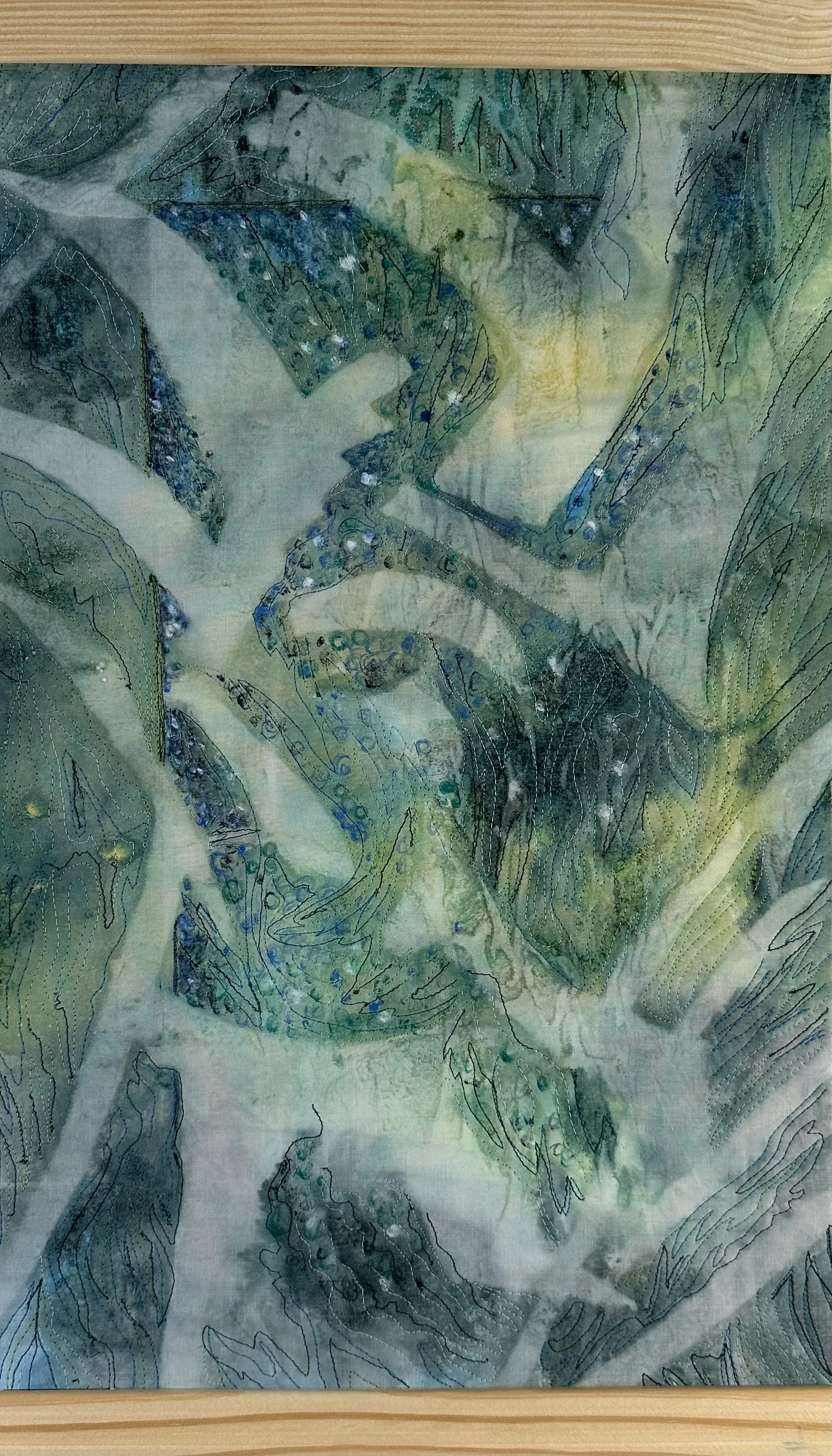 A textile art piece with abstract leaf and natural shapes in shades of green, blue, and yellow, including stitched and embroidered details on a fabric background.