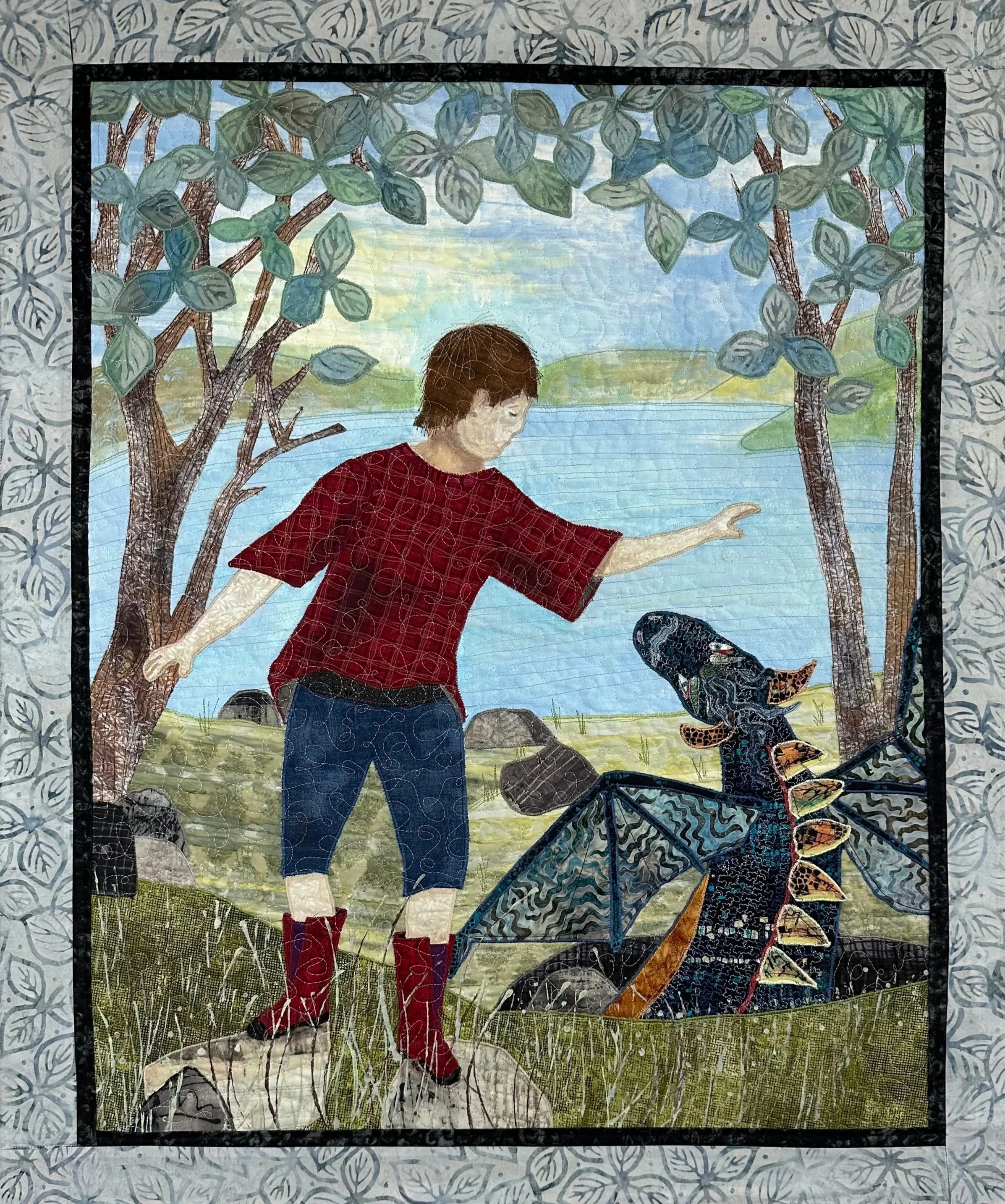 A quilted artwork depicting a person in a red plaid shirt and blue jeans standing outdoors on a grassy area by a body of water, surrounded by trees with green leaves, and a butterfly nearby.