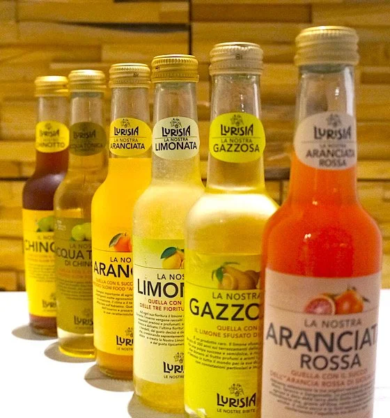 Verschillende flesjes met frisdrank, met labels zoals 'La Nostra Ranciata', 'La Nostra Limoniata', 'La Nostra Gazzosa', en 'La Nostra Aranciata Rossa', op een houten achtergrond.