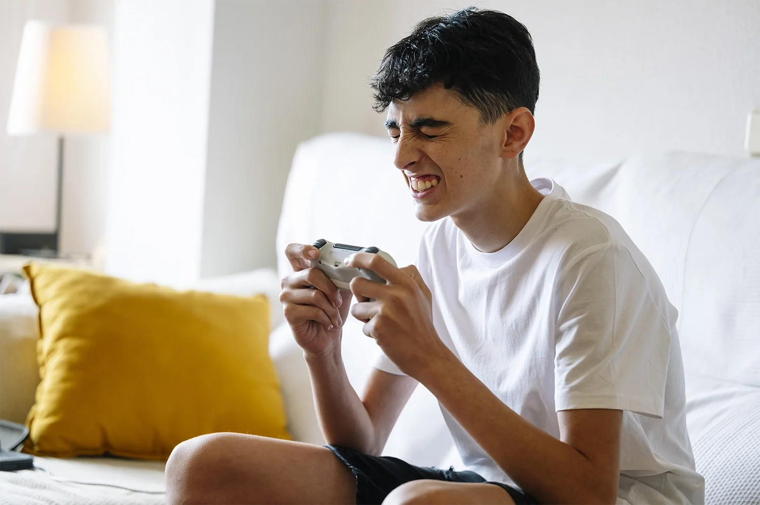 A young man sitting on a bed, smiling and holding a video game controller, engaging in gaming.