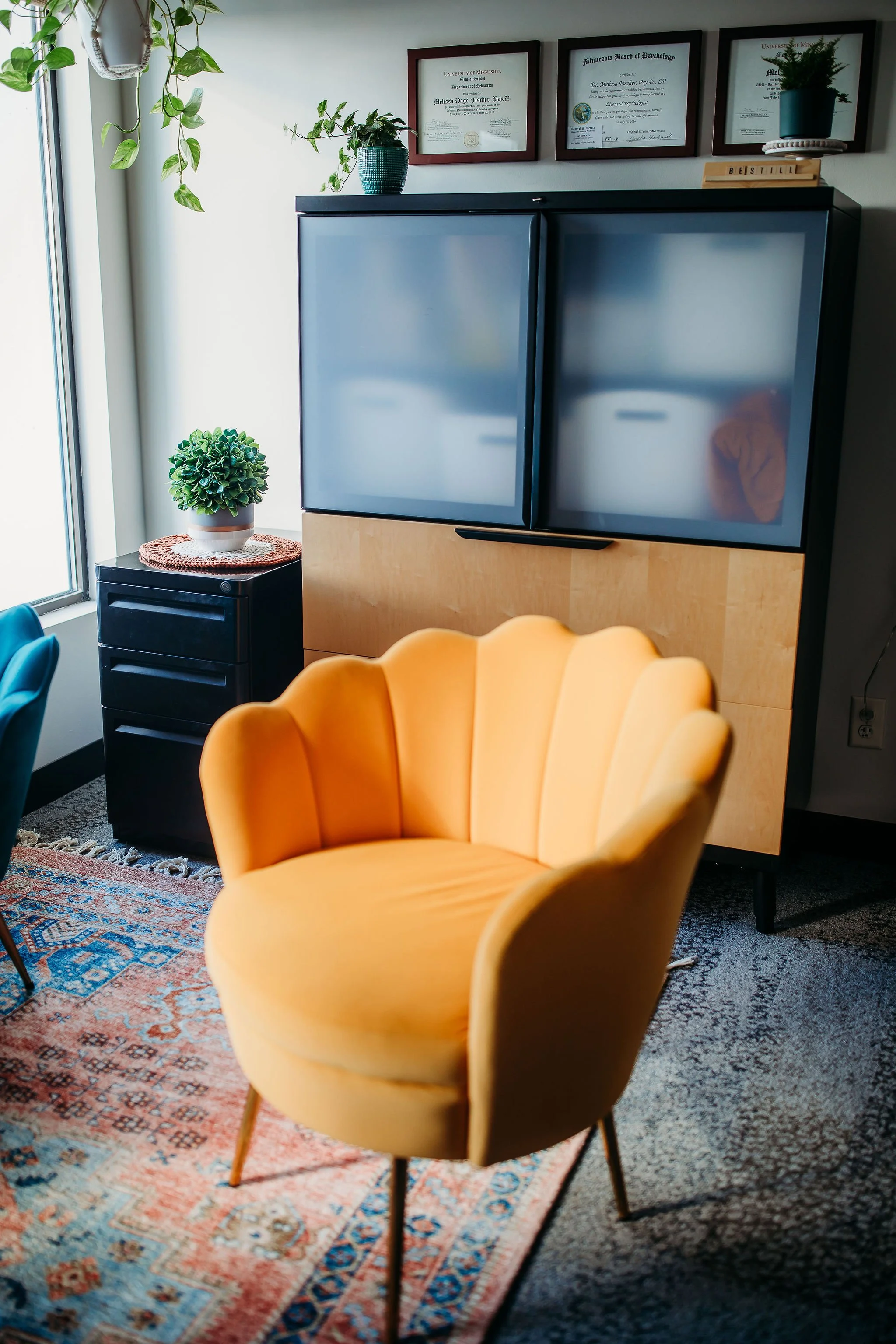 An eye-catching yellow upholstered chair with a scalloped backrest in an office or living room. Behind it is a patterned rug on the floor, a small black cabinet with a green potted plant on top, and a larger cabinet with frosted glass doors. The wall features three framed diplomas or certificates, and several potted plants are on the windowsill and cabinets.