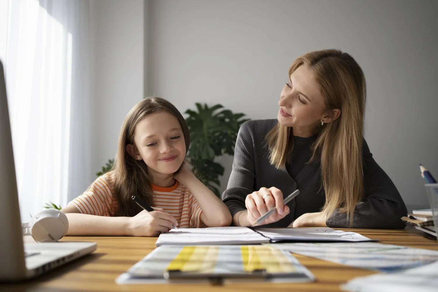 A woman and a young girl sitting at a desk together, smiling and looking at each other. The girl is holding a pen and appears to be doing schoolwork, with open notebooks and books on the desk. The woman is holding a pen and pointing to something in the notebook, with a laptop, a cup with pens, and some colored swatches also on the desk. There is a plant in the background.