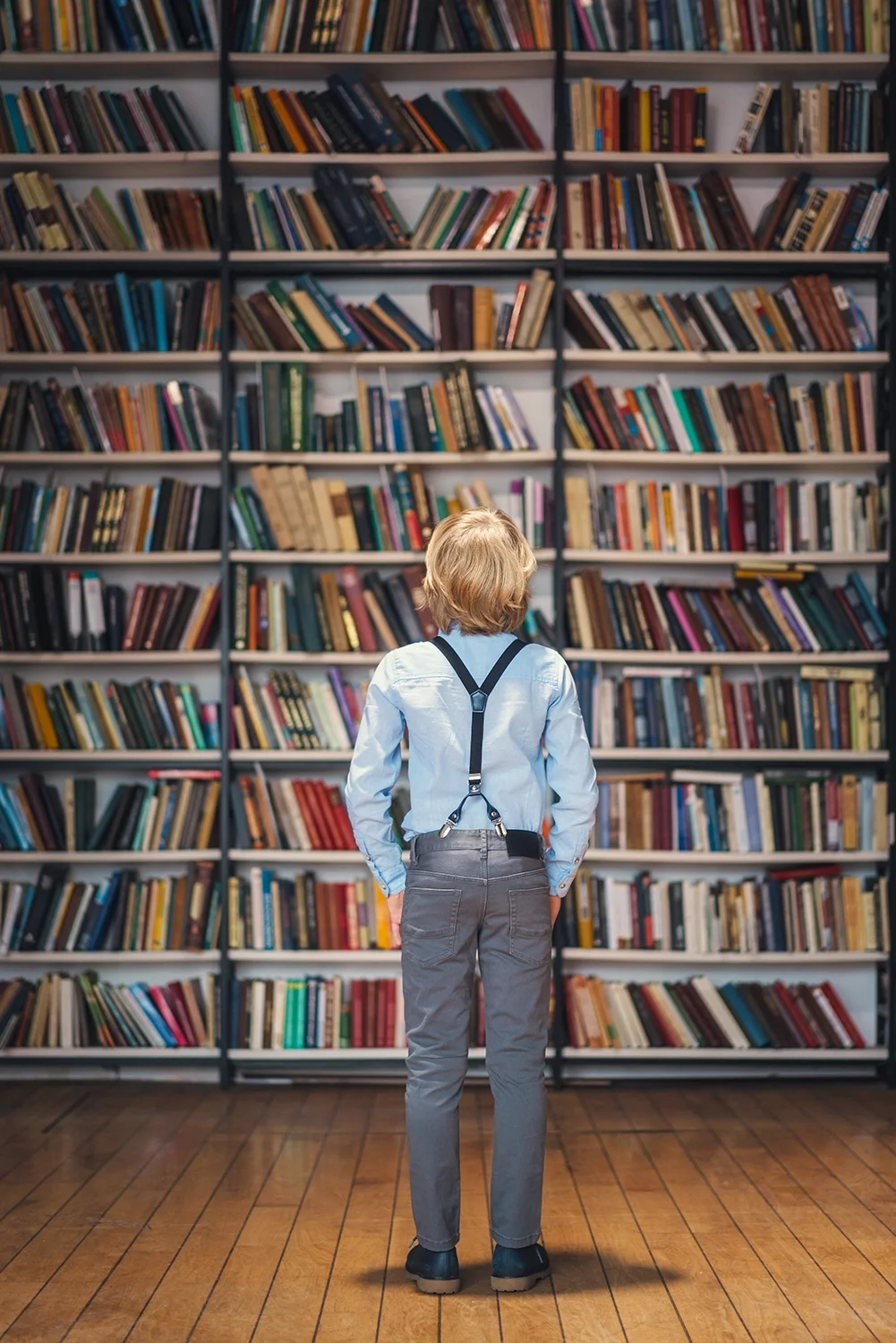 A young boy with blonde hair, wearing a light blue shirt, suspenders, gray pants, and black shoes, stands in front of tall bookshelves filled with colorful books in a library, facing the shelves.