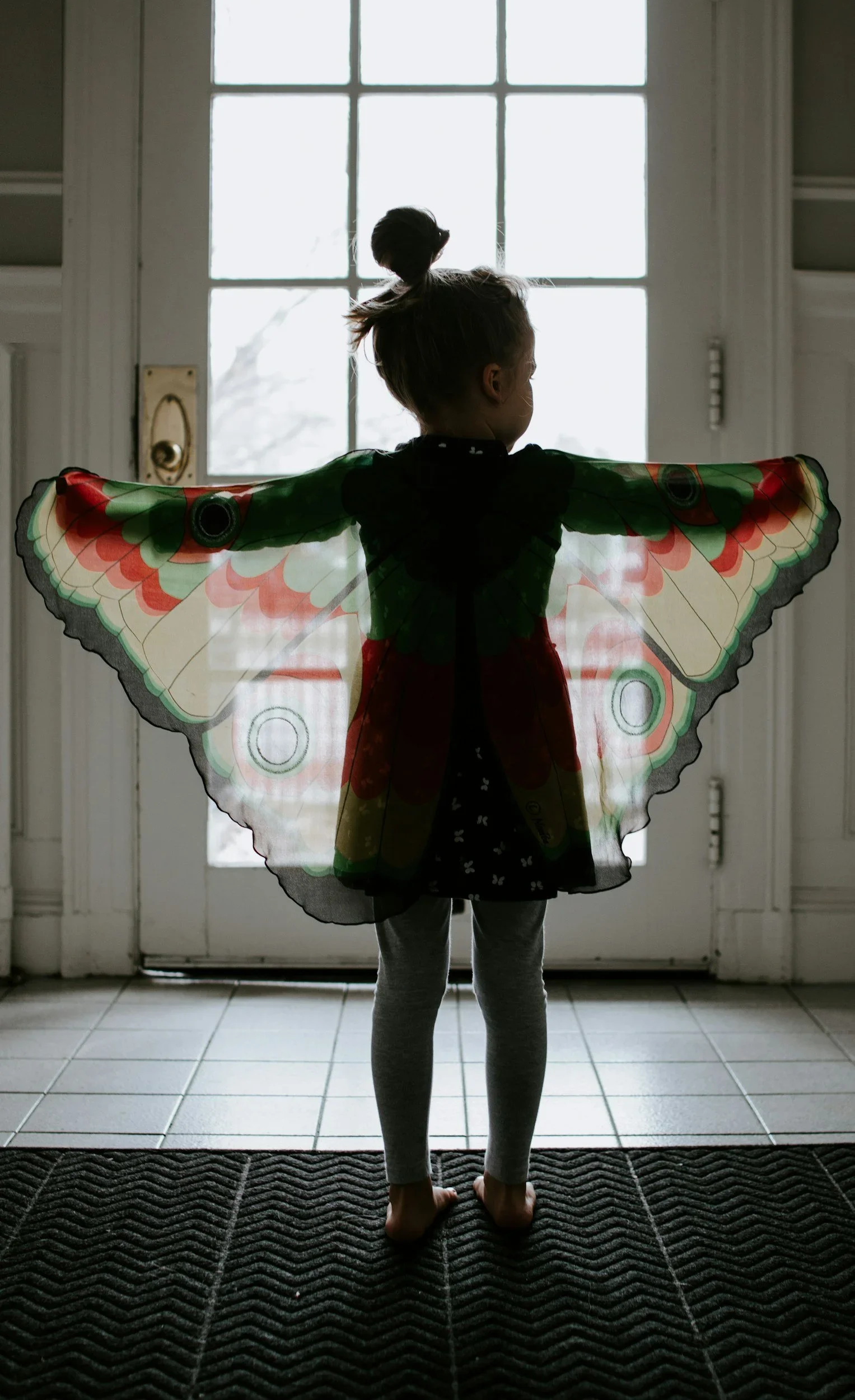 A young girl standing in front of a glass door, wearing butterfly wings and looking outside.