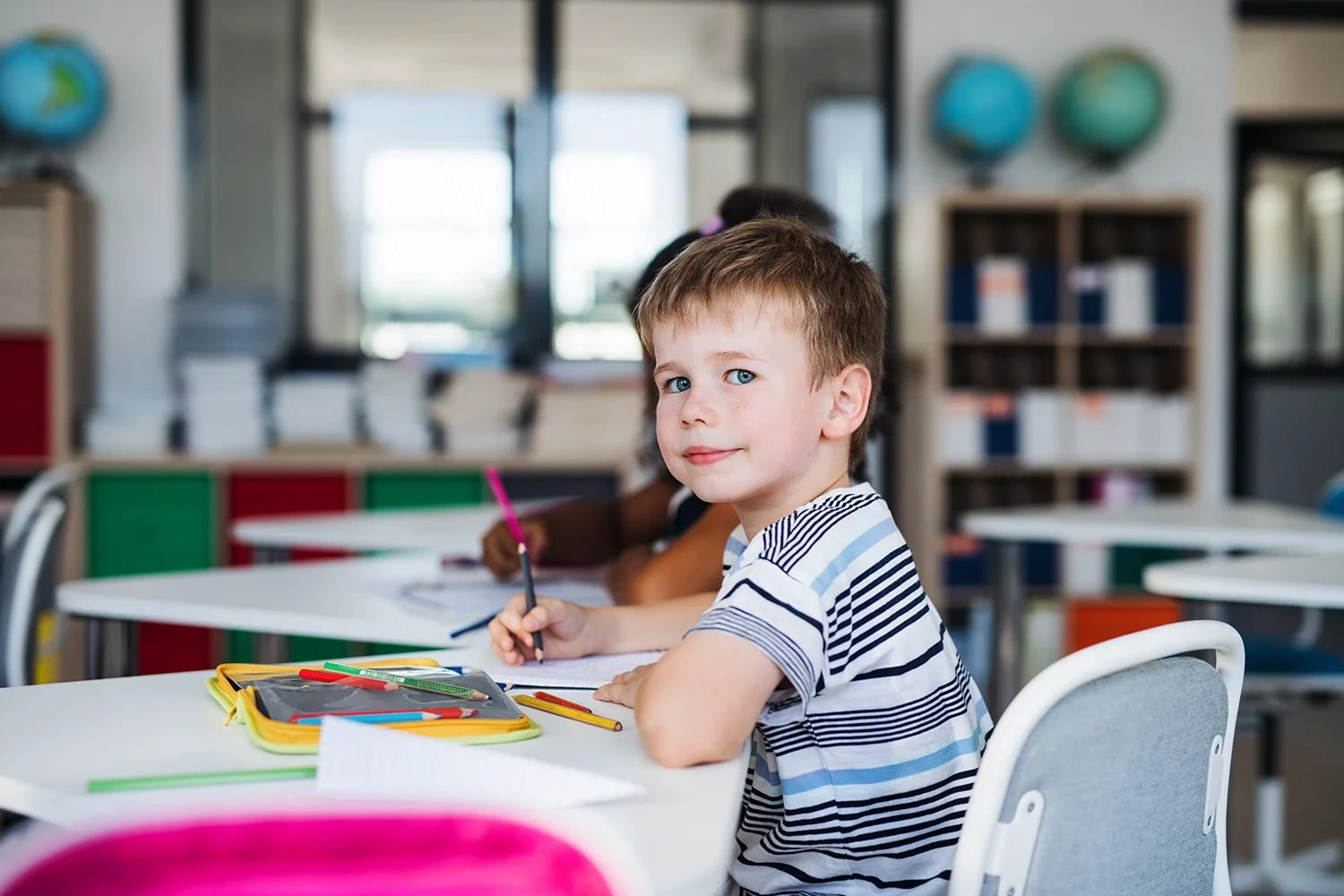 A young boy with brown hair and blue eyes sitting at a desk in a classroom, looking at the camera with a slight smile. He is holding a pencil and has a yellow pencil case with colored markers and pencils on the desk.