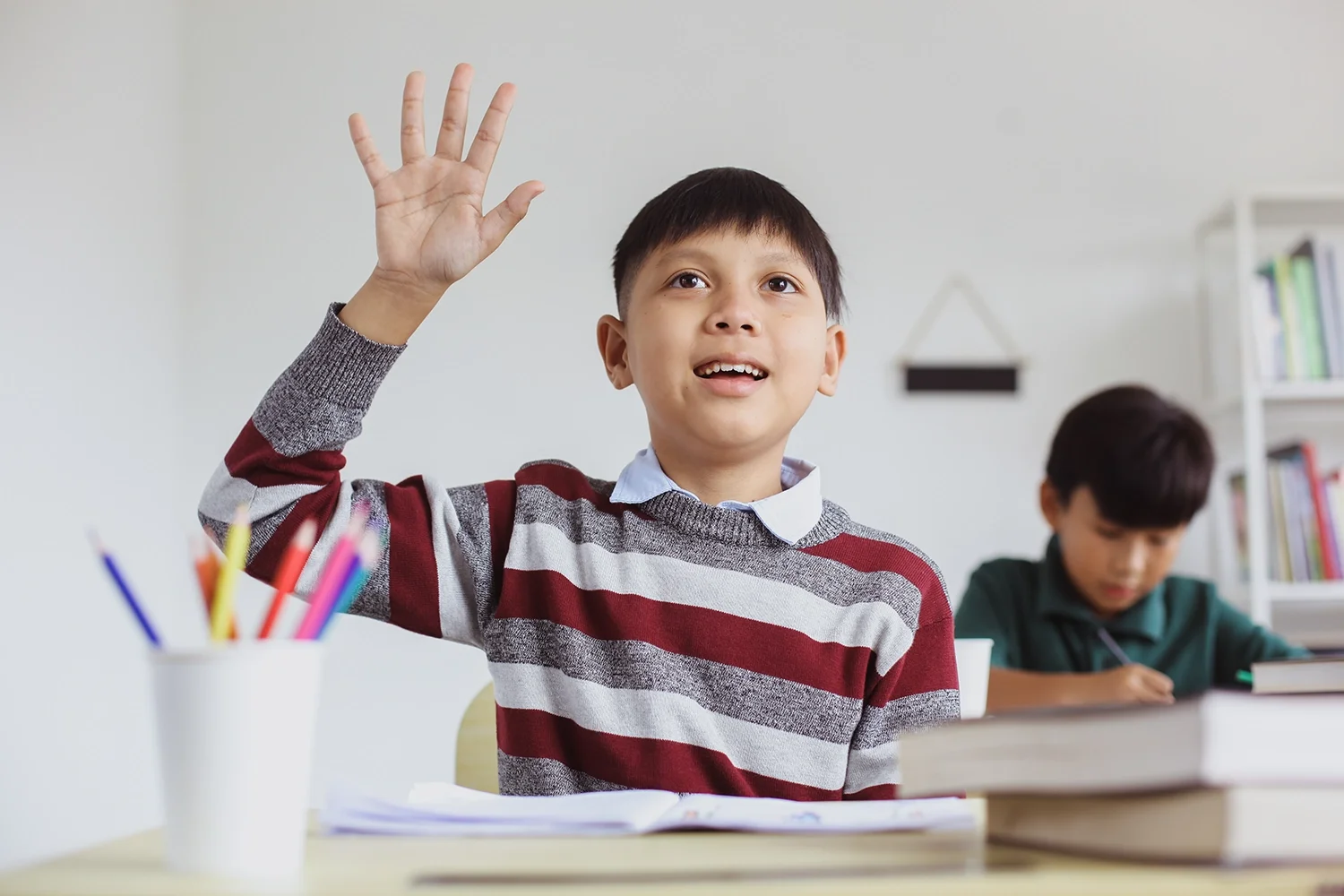 A young boy with short dark hair wearing a striped red, gray, and white sweater, raising his hand in a classroom. Another student is seated in the background, focused on his work.