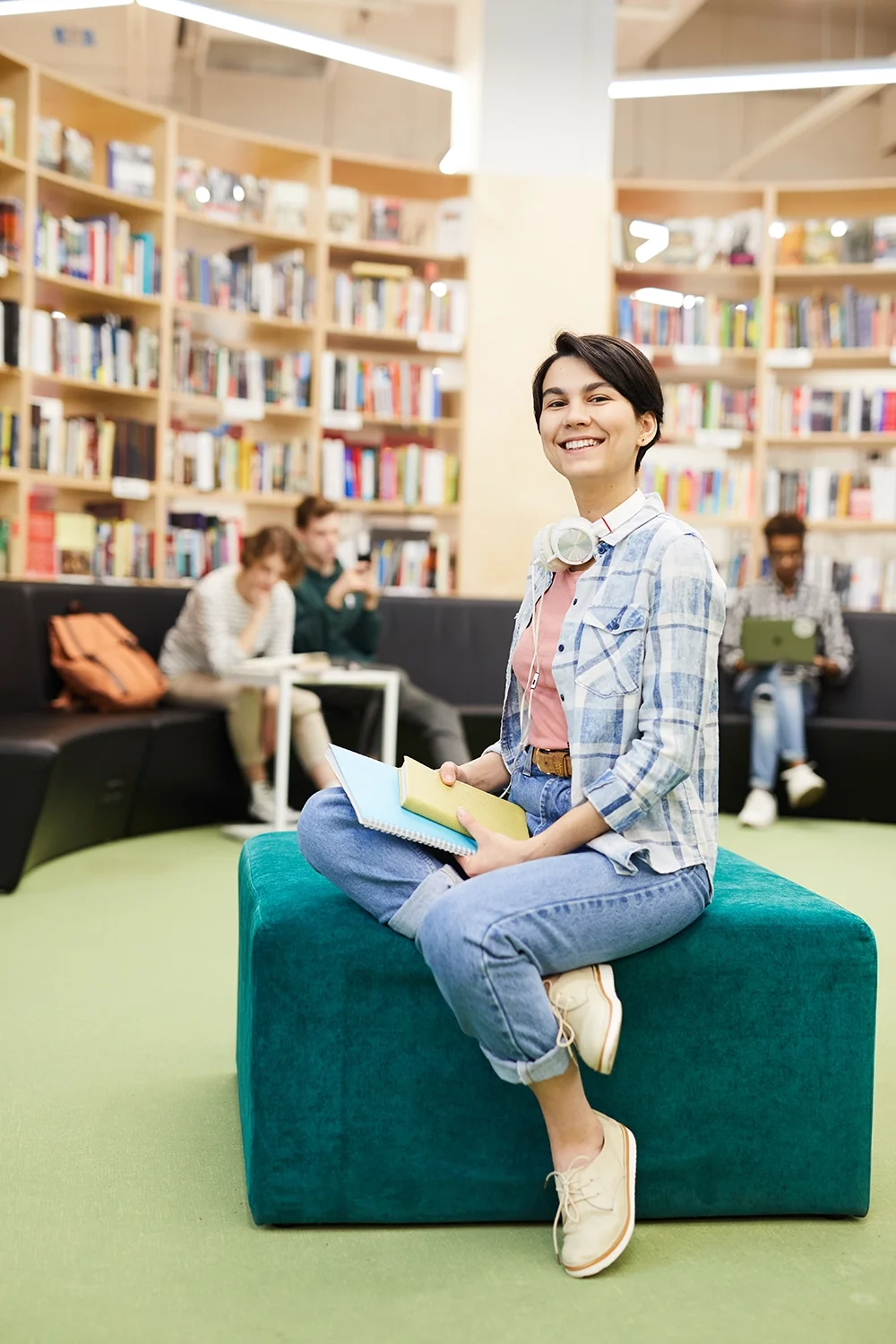 A young woman sitting on a teal ottoman in a library, smiling while holding notebooks. Behind her, there are three people seated on a black couch reading or using devices, with bookshelves filled with books in the background.