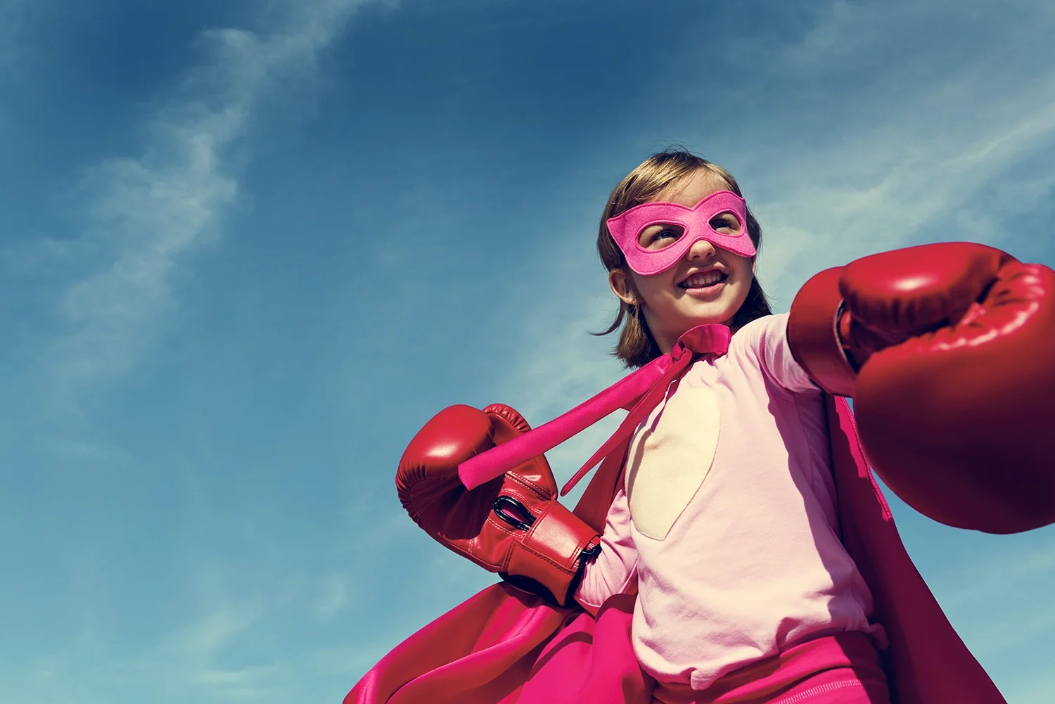 A young girl dressed as a superhero, wearing a pink mask, pink gloves, and a pink cape, poses with her fists up against a blue sky with some clouds.