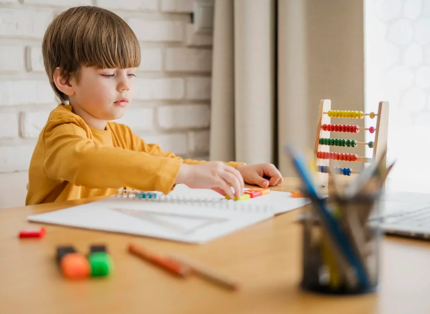 A young boy in a mustard yellow shirt playing with colorful educational toys on a white sheet at a desk, with an abacus and art supplies nearby, in a well-lit room with a white brick wall and a window in the background.