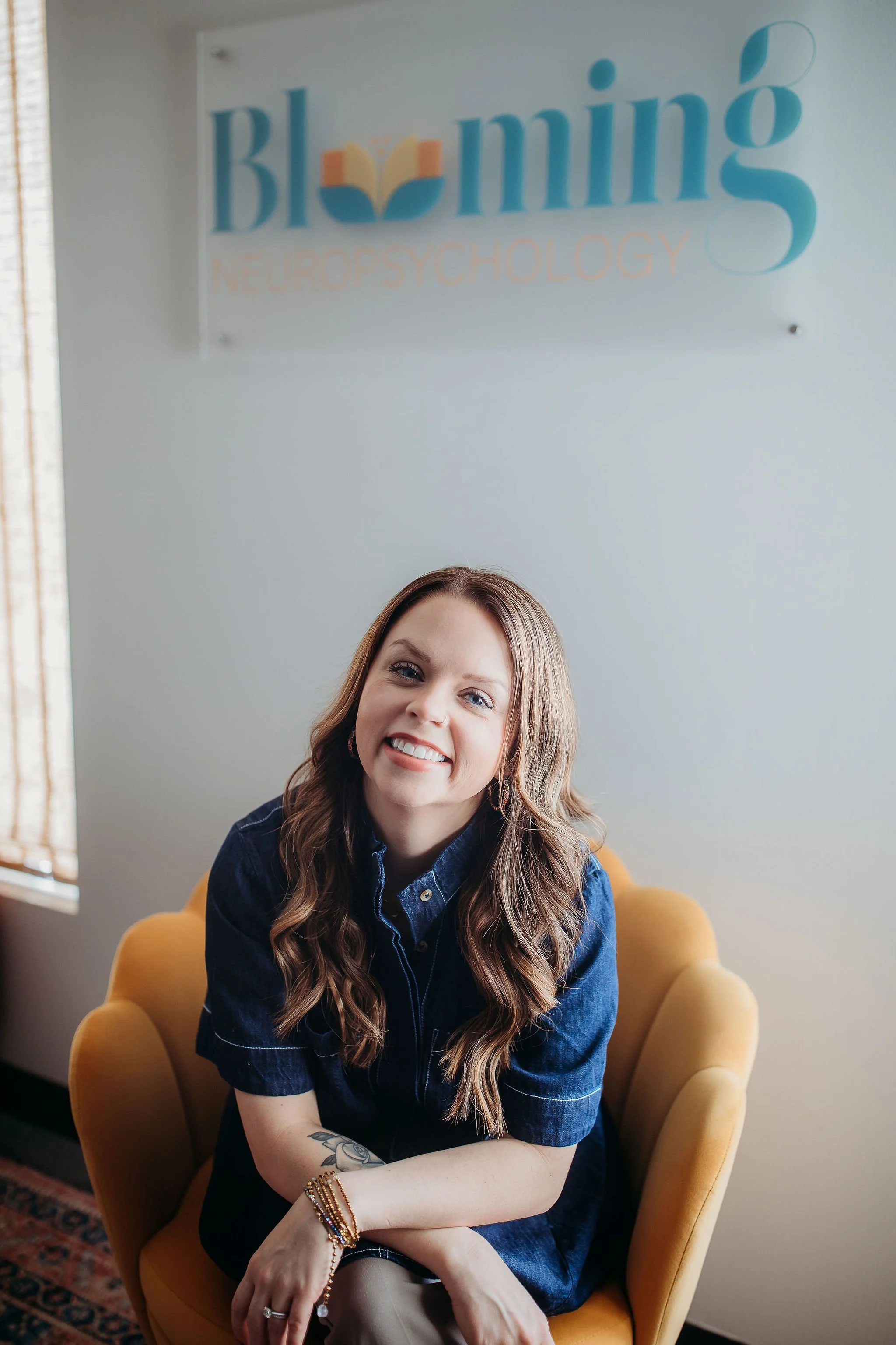 A woman with long, wavy brown hair sitting in a yellow armchair, smiling, in front of a blurred sign that reads 'Bloom Neuropsychology' on the wall.