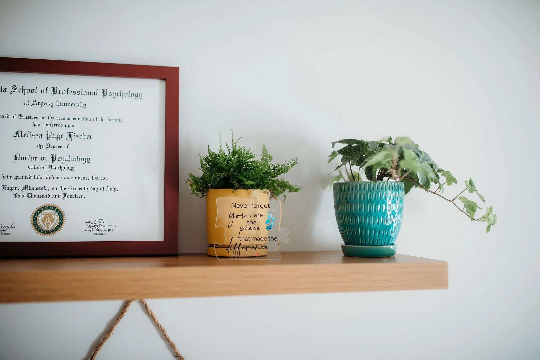 A wooden shelf holding a framed diploma from Argosy University, a potted green fern, and a potted ivy plant on a white wall background.