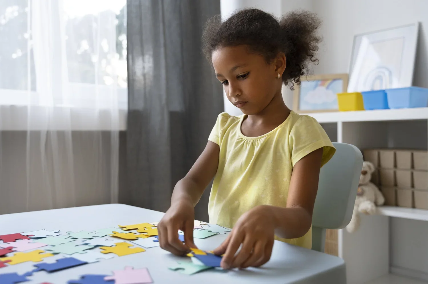 A young girl with curly hair, wearing a yellow shirt, assembling colorful puzzle pieces at a white table in a well-lit room.
