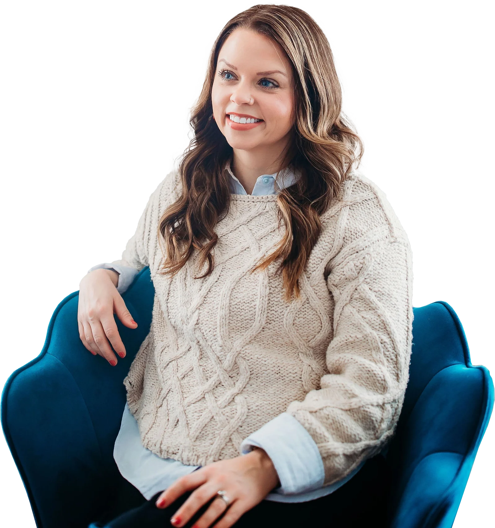 A woman with long, wavy brown hair and a fair complexion sitting in a blue armchair, smiling, wearing a cream cable-knit sweater over a white collared shirt.