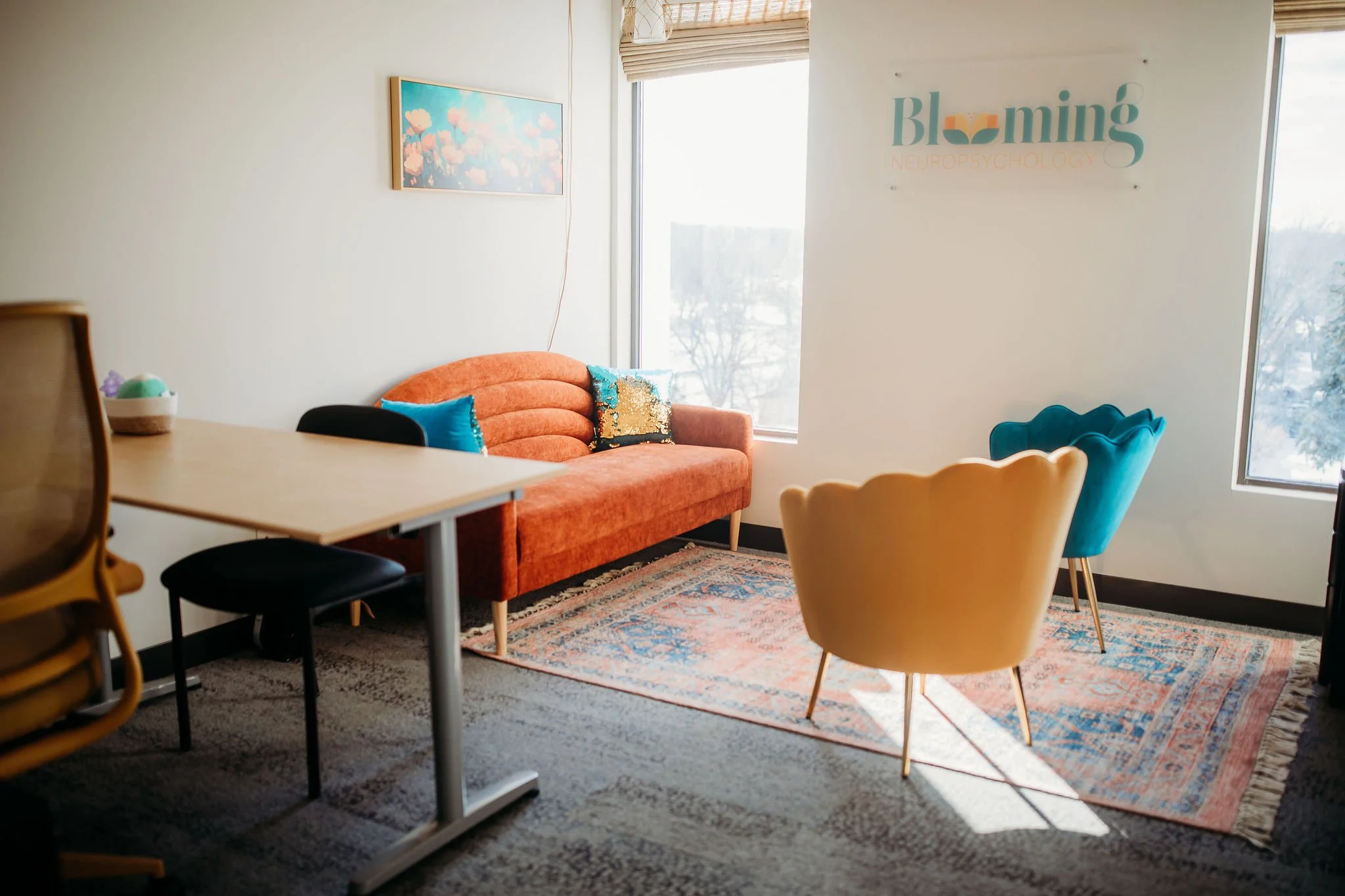 Modern office reception area with orange velvet sofa, two yellow and two blue accent chairs, a patterned rug, a wall art and a large window.
