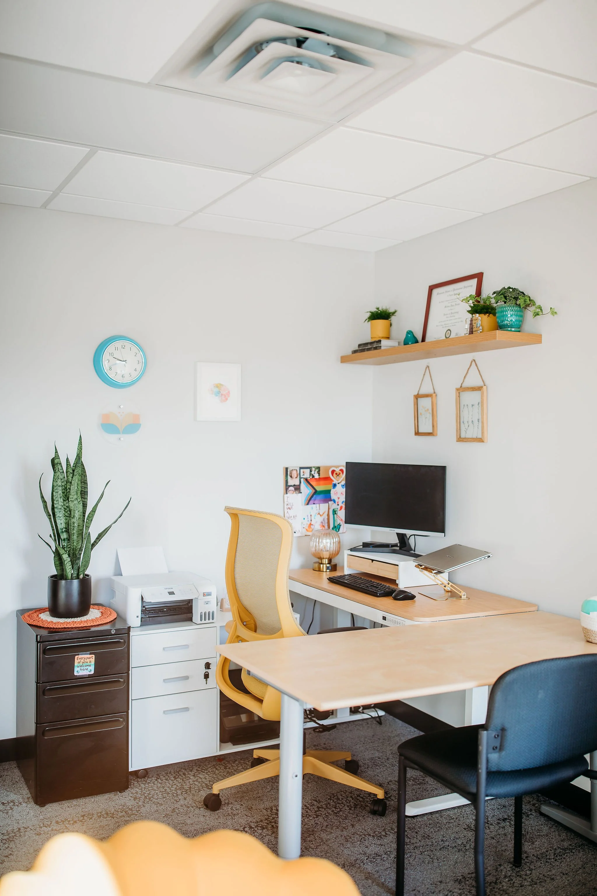 A home office with a yellow ergonomic chair, a black office chair, a desk with a computer monitor, keyboard, and desk lamp, a bookshelf, some wall art, a potted plant, and a ceiling air vent.